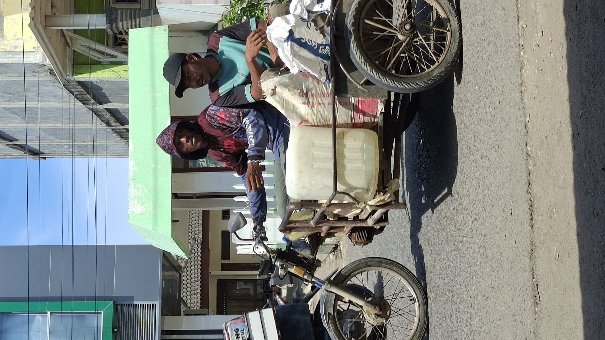 Two young men are riding on a motorized three-wheeled vehicle on a paved road in Lhokseumawe, Indonesia. The vehicle has a trailer attached to the back, carrying a large white plastic container and several large sacks. The man in the front is wearing a maroon hoodie with intricate patterns and a patterned hood pulled up. The man in the back is wearing a teal and black t-shirt and a black baseball cap. They are both looking forward, seemingly engaged in their journey. The background shows buildings with various architectural styles, including a modern building with a green facade and a taller, older building. The sky is clear and blue, indicating a sunny day. The road surface appears to be asphalt with some minor cracks and debris. The overall scene depicts everyday life and transportation in an urban environment.