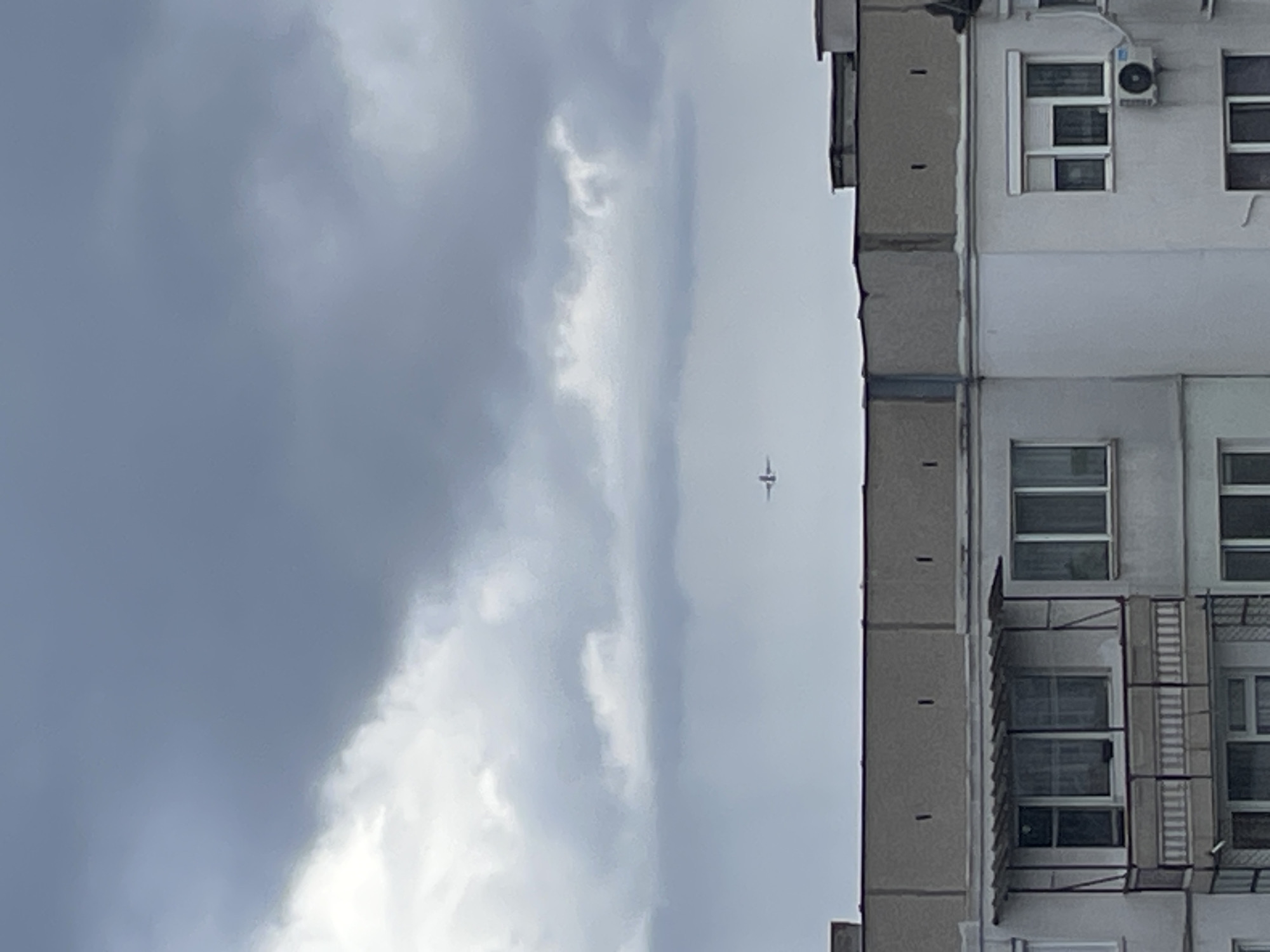 The image depicts a cloudy sky in Sofia, Bulgaria, with a plane visible in the distance. Part of a grey concrete building is in the foreground to the right. The building has a visible fire escape. The sky is grey with patches of white clouds.