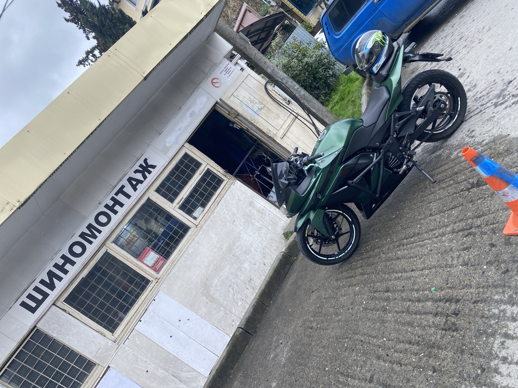 The image shows a green Kawasaki motorcycle parked on a paved street next to a building with the Russian word "ШИНОМОНТАЖ" displayed prominently, which translates to "tire fitting" or "tire service". The building has a beige exterior with gridded windows and a white door. A blue truck is parked partially visible in the background. A construction cone is positioned near the motorcycle on the right side of the image. The setting appears to be an urban area, possibly Sochi, Russia. The sky is overcast.