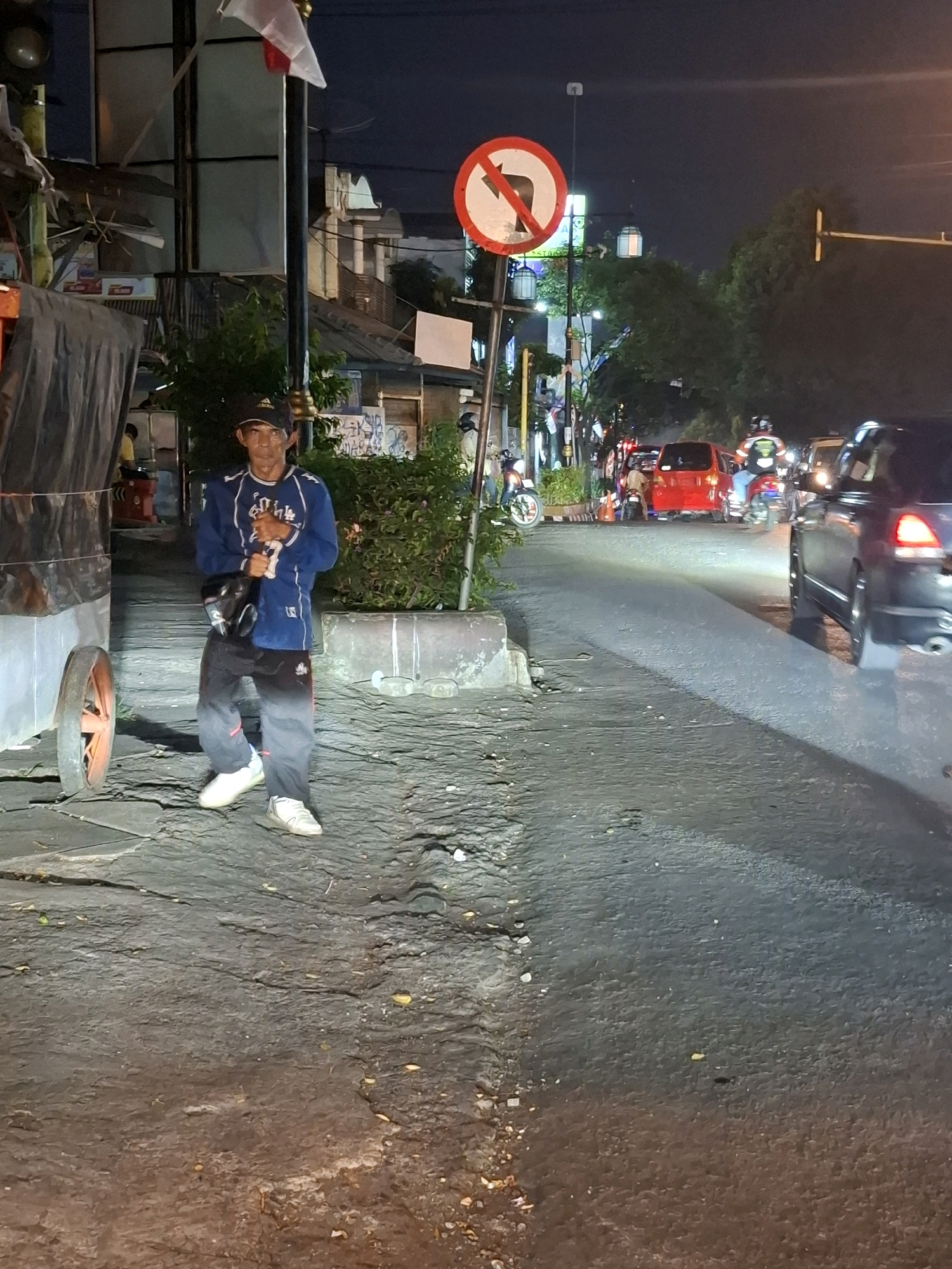 The image shows a street scene at night in Cianjur, Indonesia. In the foreground, a man is standing on a cracked pavement. He is wearing a dark baseball cap, a blue athletic jacket with white and red accents, and dark, gradient pants. He has a dark bag slung across his chest. To the left of the man is a large, weathered cart with a partially visible orange wheel. Behind the man, a dense green bush is present.
A "no right turn" traffic sign is visible in the mid-ground, mounted on a pole with a white flag attached to its top. The street is lined with various structures and trees. Traffic, including cars and motorcycles, is visible on the road, with headlights and taillights illuminating the scene. The lighting suggests it is nighttime, possibly after dusk. The overall environment appears to be an urban or semi-urban street.