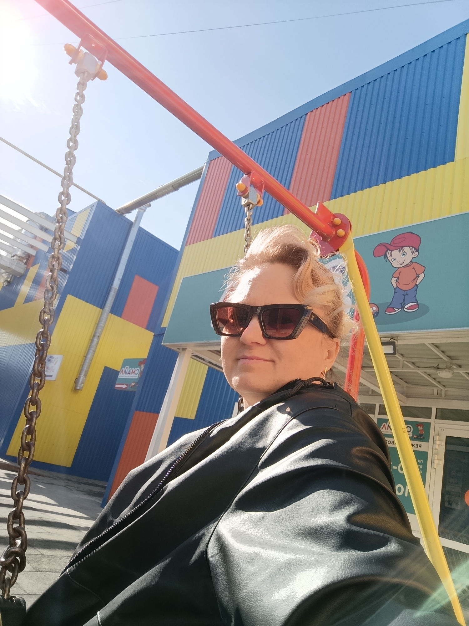 An adult female, likely middle-aged, is depicted outdoors in a sunny daytime scene, seated on a swing. She wears dark-framed sunglasses and a black faux leather jacket with prominent zippered sleeves. Her blonde hair is styled in a wave, and a small gold hoop earring is visible on her left ear. A metal chain from the swing hangs vertically on the left, while a red horizontal bar and a yellow diagonal support pole, part of the swing structure, are visible above and to her right.
Behind the woman is a commercial building with a brightly colored facade constructed from corrugated metal panels. These panels are arranged in large, irregular blocks of blue, yellow, and red/orange. On the upper right, a light blue section of the facade features a cartoon illustration of a boy in a red cap and shirt, standing next to a small red toy car. Below this illustration, a glass entrance reveals inverted Russian text, including "АНАМО" (ANAMO), indicating a children's store. The specific location is identified as outside the ANAMO children's store at Vokzalnaya Ulitsa, 1/1, Chaikovsky, Perm Krai, Russia. Bright sunlight from the upper left creates lens flare and casts distinct shadows on the ground visible on the left.