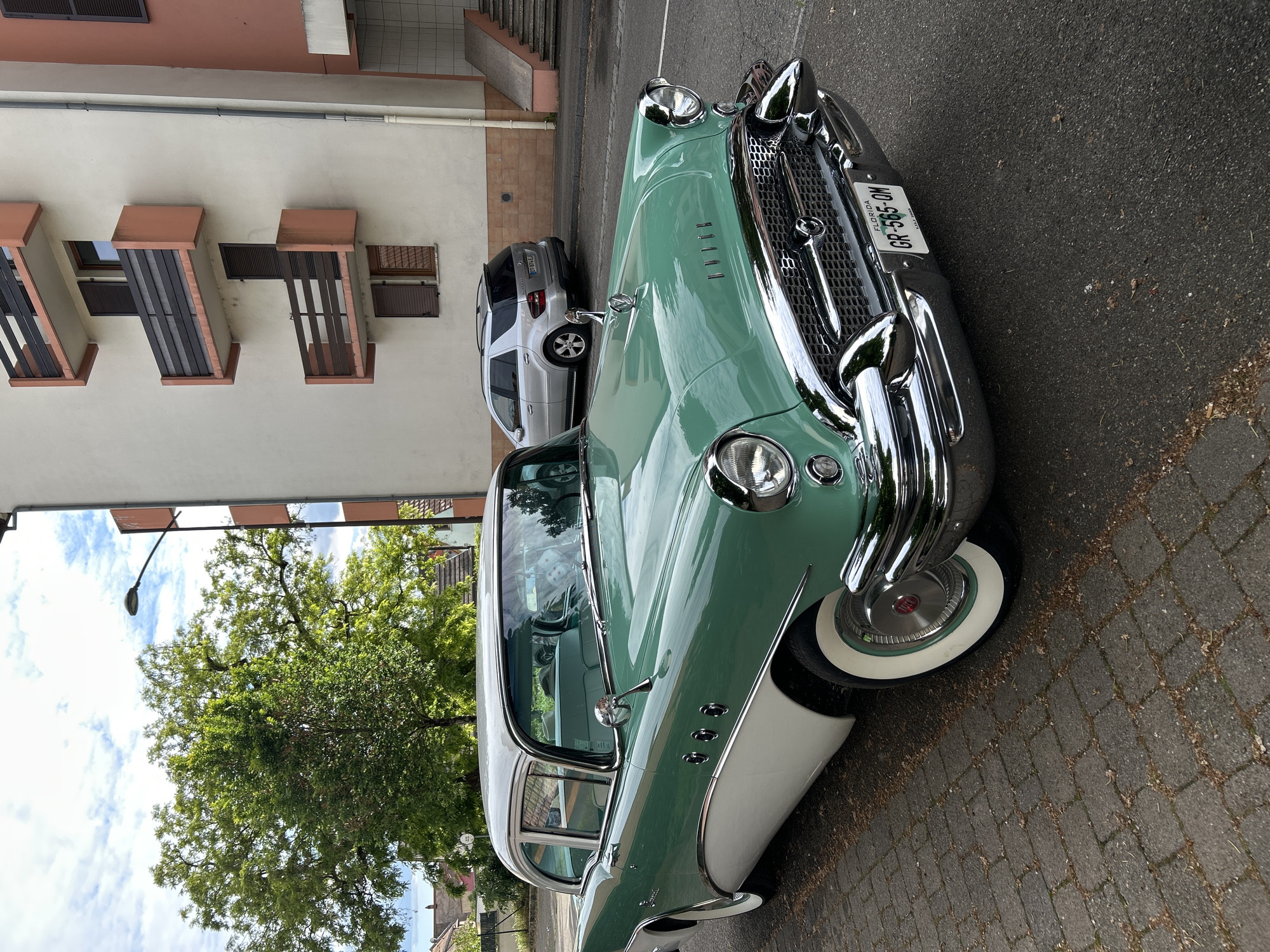 The image depicts a vintage light green and white Buick automobile parked on a cobblestone street. The license plate on the car reads "FLORIDA GR-565-DM." Adjacent to the car is a modern silver SUV, and behind them is a building with distinctive protruding window features, suggesting European architectural style. A large tree is visible in the background, its leaves partially obscuring the building behind. The setting appears to be a residential street in Strasbourg, France. The photograph seems to have been taken at street level.