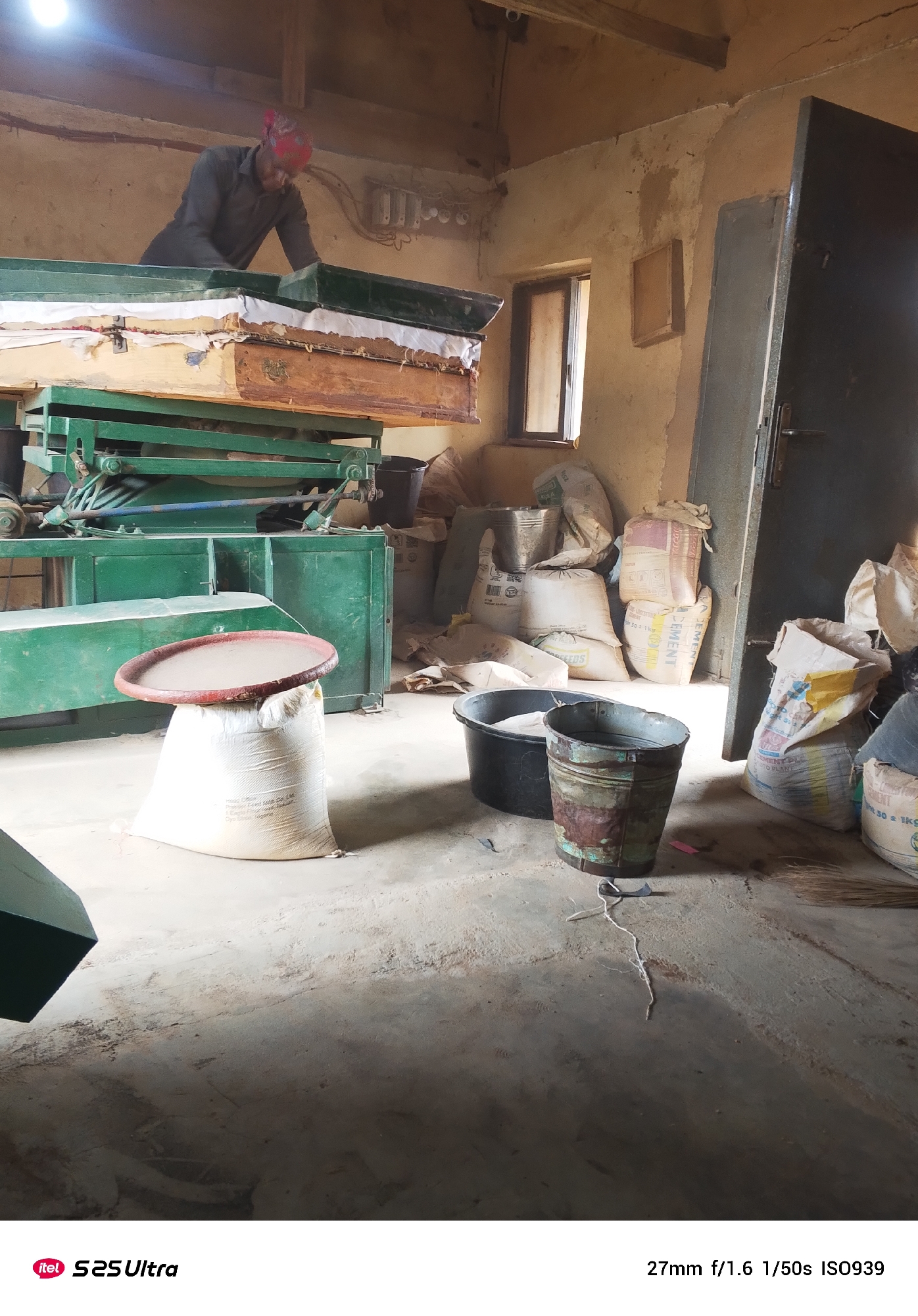 An indoor shot shows a person wearing a red headscarf and a dark shirt working with a machine. In the foreground, a large white bag of what appears to be cement is topped with a red, round tray. Behind it, two buckets sit on the floor, one black and one metal with rust and paint damage. The room is cluttered with sacks of cement and other materials, with a large metal door on the right side of the frame. The walls are rough and unfinished, suggesting a workshop or storage area.