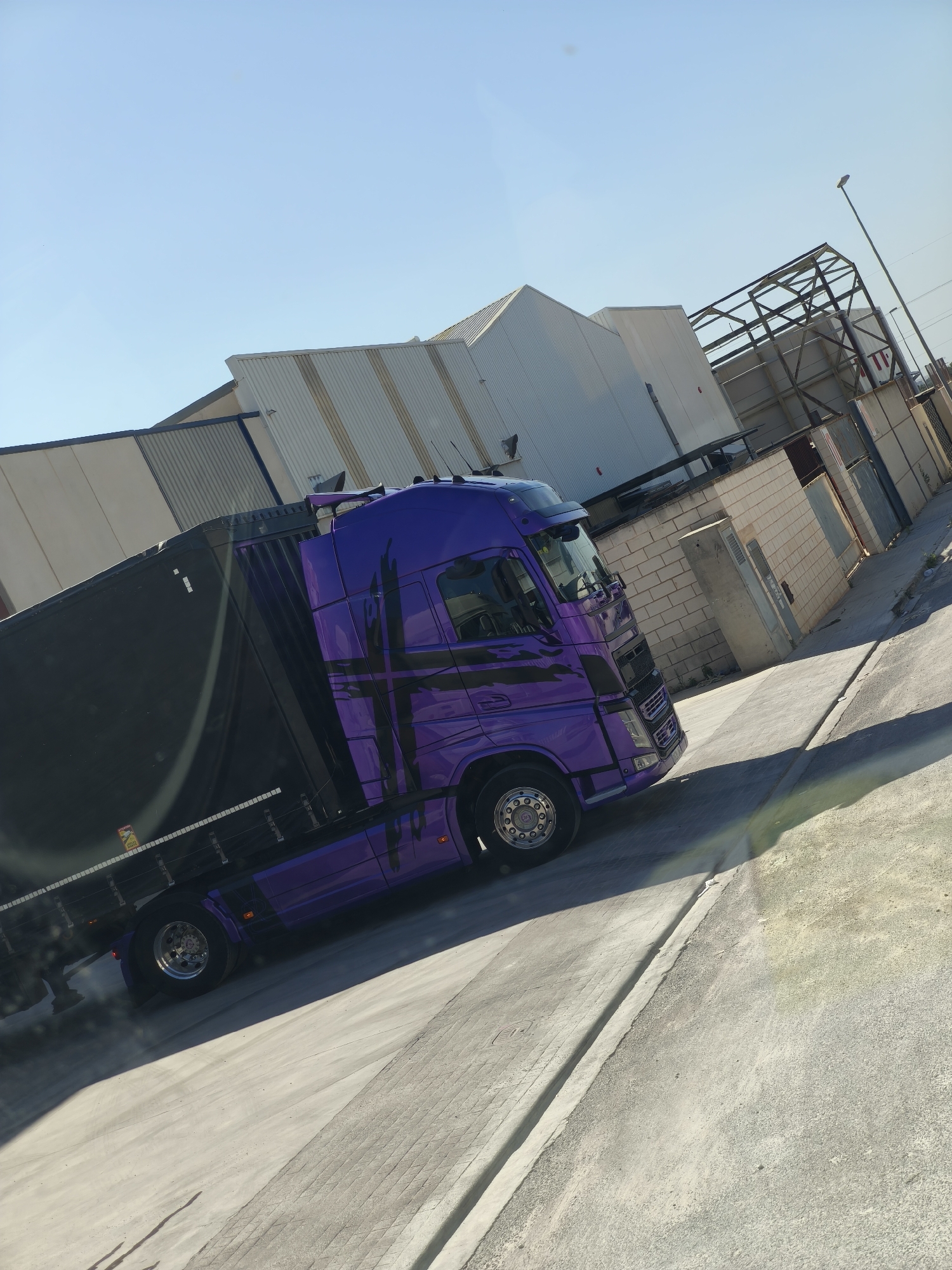 A vibrant purple semi-truck with a black canvas-covered trailer is the central subject of this image, captured in an industrial park within Polígon Industrial Dels Vents, Spain. The truck's cab features a distinctive black abstract graphic design and polished chrome wheels.
The setting consists of various industrial buildings, primarily featuring corrugated metal siding in shades of grey and white, under a clear, bright blue sky. Some structures appear to be warehouses or factory units. In the background, there is also the skeletal metal framework of what might be a partially constructed or deconstructed building or sign, and a tall street light pole. Lower walls constructed from light-colored brick are also visible, along with concrete utility boxes and other industrial elements.
The truck is situated on a paved asphalt and concrete lot or road, with strong, defined shadows cast by objects, indicating bright sunlight and a clear day, likely during mid-morning or afternoon. There isn't clear activity, but the truck appears to be parked or moving slowly within the industrial environment.
A notable detail is a small, rectangular yellow sticker affixed to the side of the black trailer, prominently displaying the word "ATTENTION" above a black symbol and some unreadable smaller text. The image is captured from an angled perspective, giving a dynamic feel to the industrial scene.