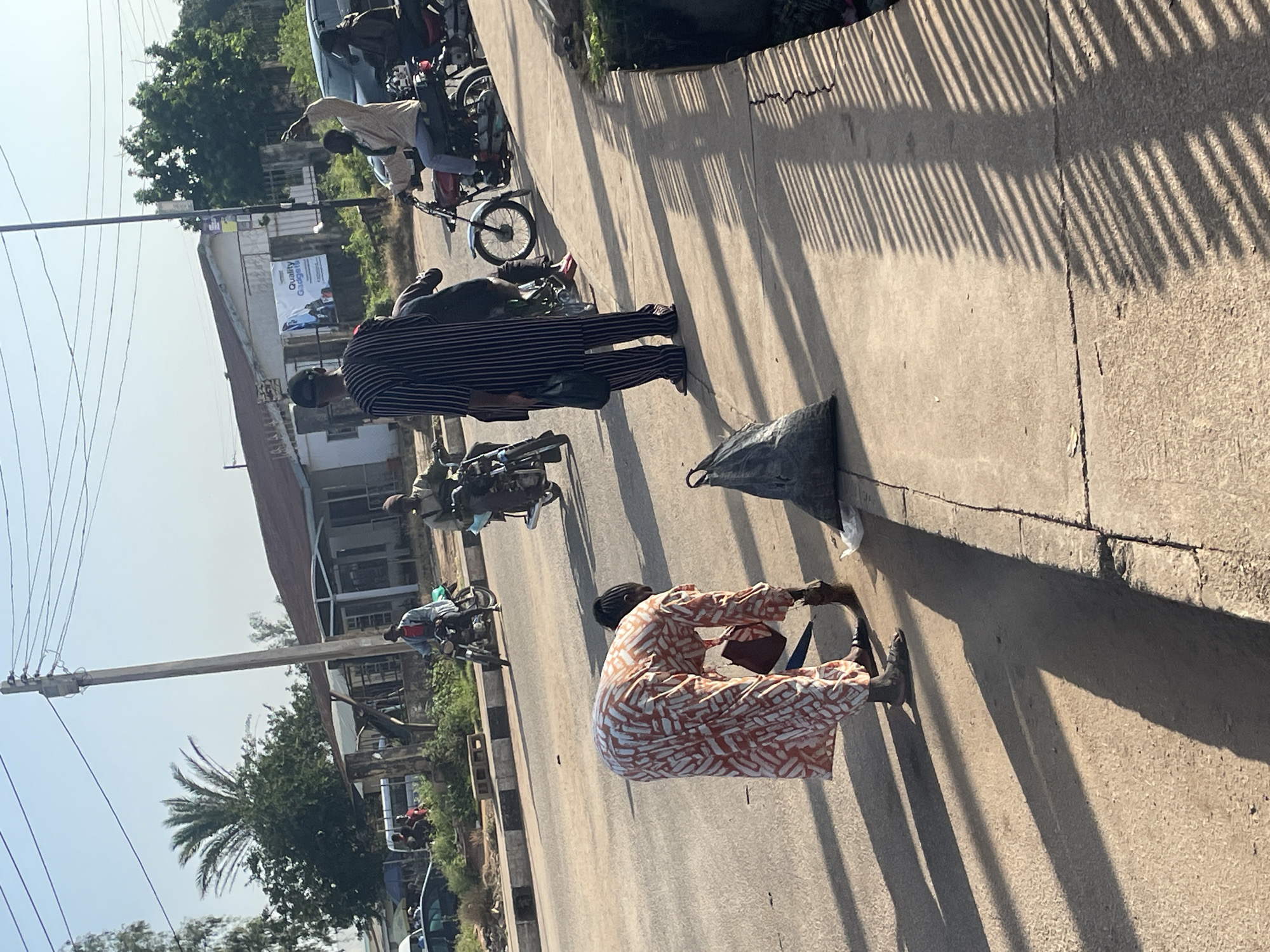 The image captures a street scene in Jos, Nigeria, during daylight. A woman in a patterned orange and white dress is bent over on a concrete sidewalk, appearing to pick something up or work on the ground. Beside her, a large black garbage bag rests on the sidewalk, and another smaller white plastic bag is visible near the curb. In the background, several people are on motorcycles, some in motion and others stationary. One man in a black and white striped outfit stands on the sidewalk. The road is paved, and there are trees and buildings lining the street. Power lines crisscross the sky, and the sun casts long shadows on the ground, suggesting it is either morning or late afternoon. The environment appears to be an urban or semi-urban area with a mix of traffic and pedestrian activity.