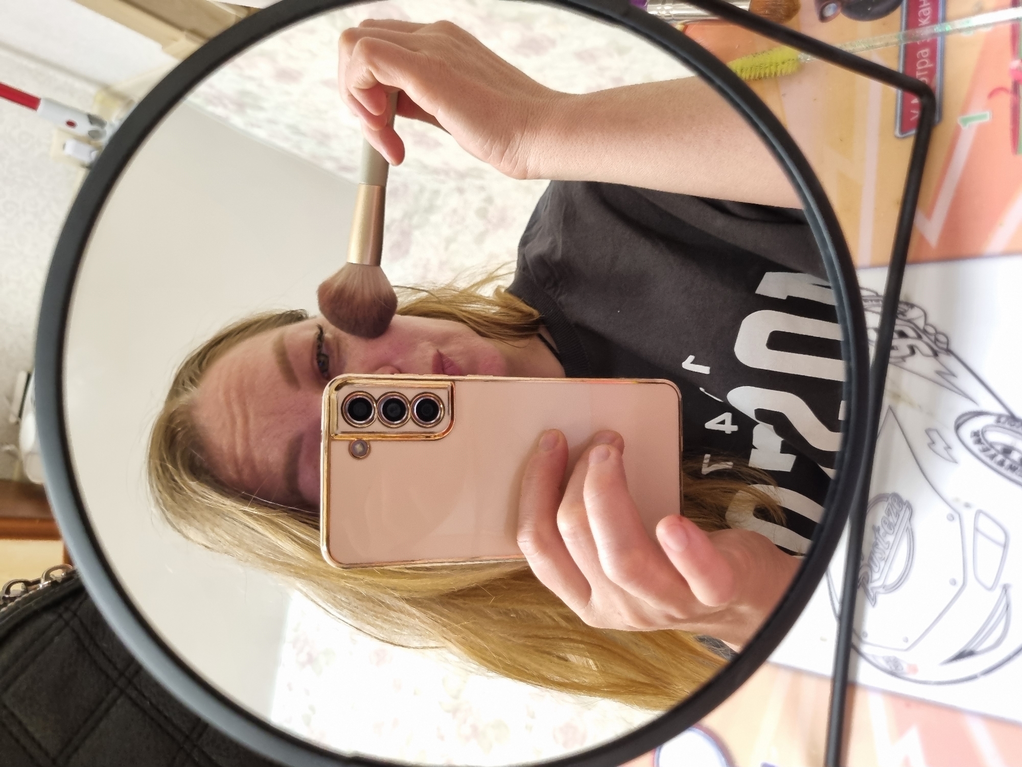A person is taking a selfie in a round black-framed mirror. The person is holding a makeup brush to their face with one hand and a pink smartphone with three camera lenses with the other. The person has blonde hair and is wearing a dark-colored shirt with white lettering. In the background, there is a light-colored wall with floral patterns. On a table in the background, there are makeup brushes and a paper with writing in Russian, potentially indicating the city of Chaikovsky, Russia. A black purse hangs on the bottom left of the mirror frame.