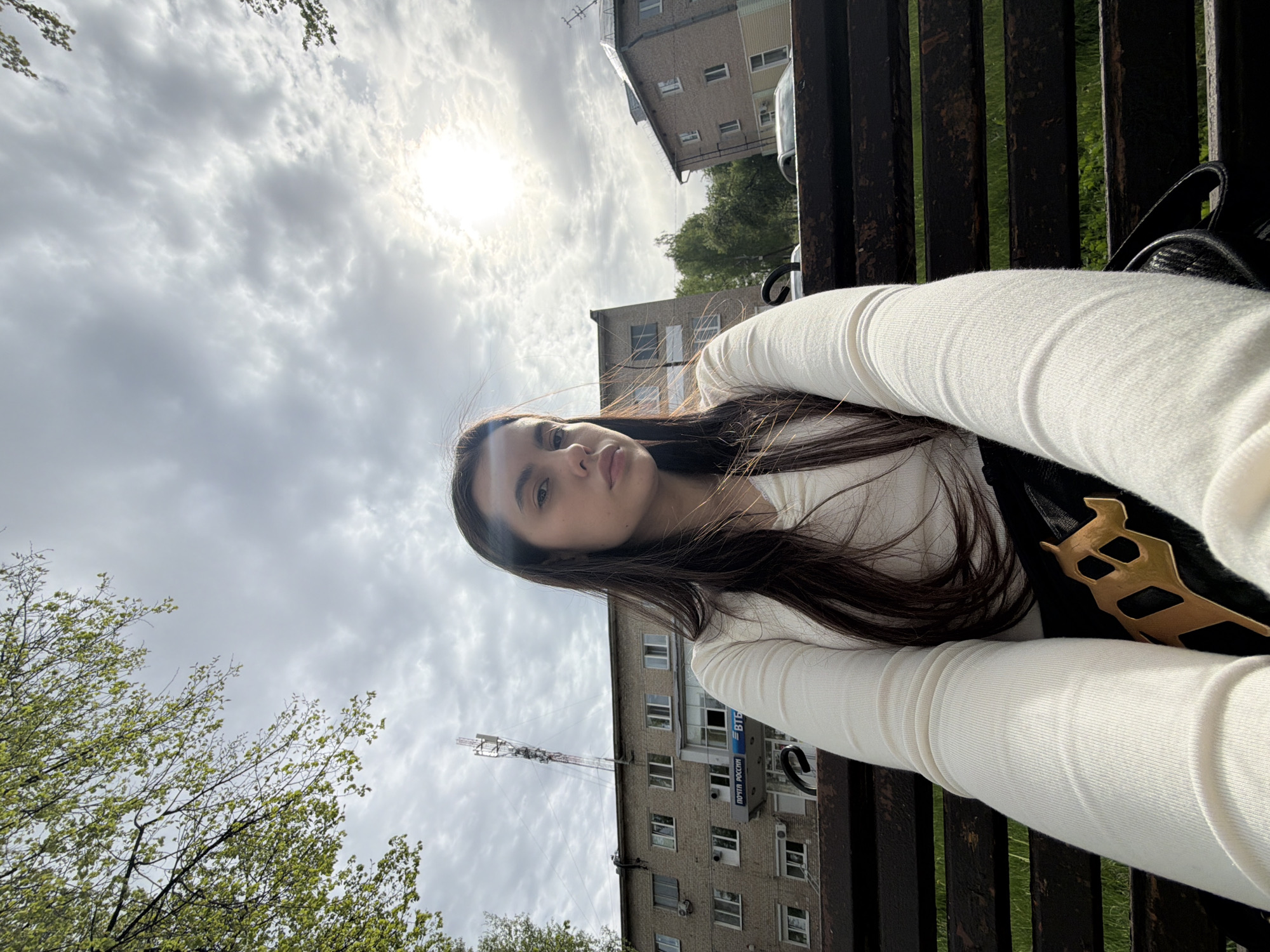A young woman with long dark hair is sitting on a park bench with her arm extended in a selfie pose. She wears a white long-sleeved shirt. The background reveals a cityscape under a cloudy sky with the sun shining through. Buildings, including one with a postal service sign in Russian ("Почта России" - Post of Russia), are visible, as is a communications tower. Given the presence of the postal service signage, the location appears to be the city of Chaikovsky, Russia. A backpack with a gold decorative piece rests beside her on the bench.