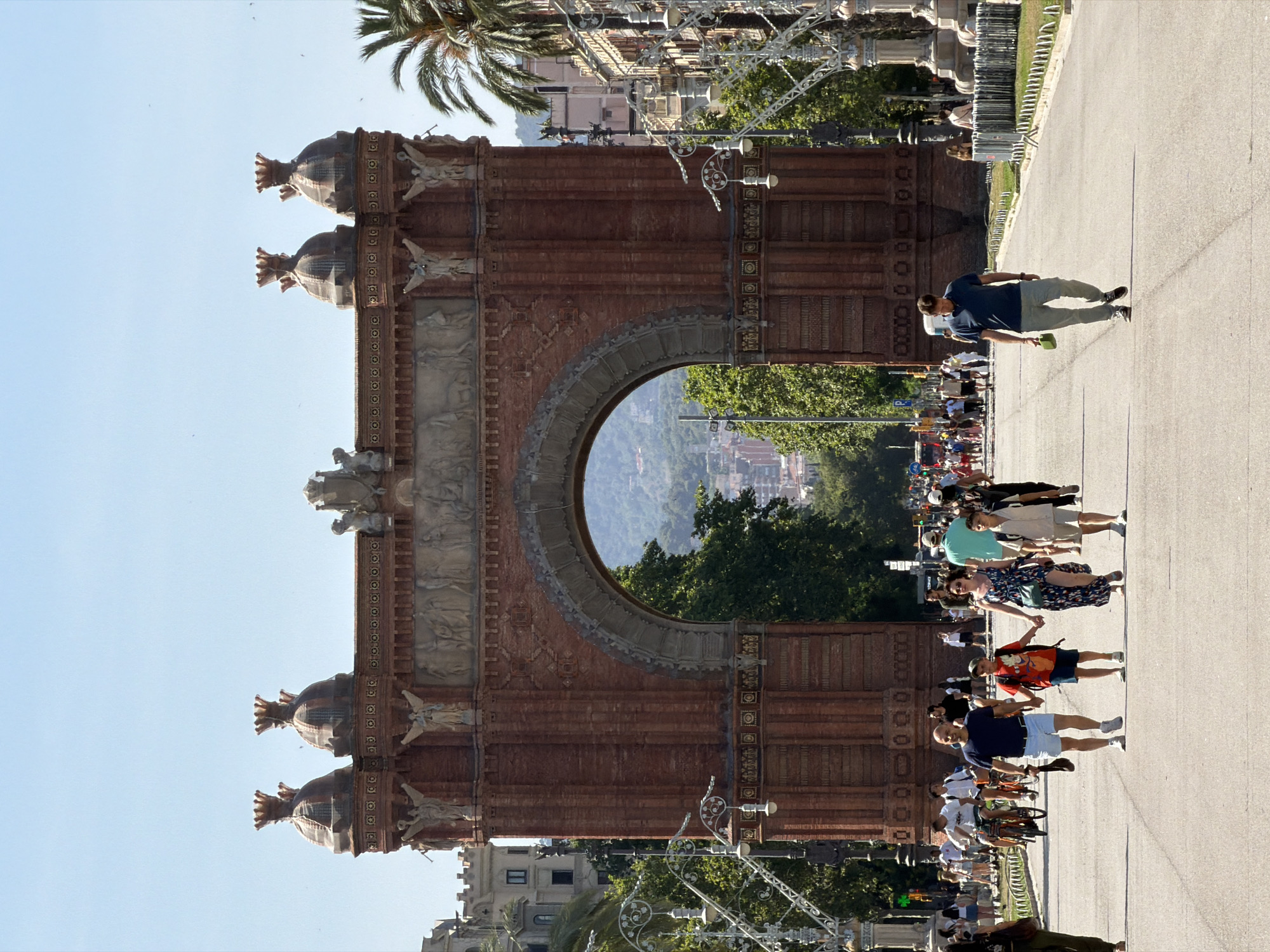 The image shows the Arc de Triomf in Barcelona, Spain, on a bright, sunny day. The arch is made of red brick and is adorned with sculptures and decorative elements. In the foreground, a paved path leads towards the arch. Several people are walking on the path, some individually and some in groups. A family of four, consisting of two adults and two children, is walking towards the camera, holding hands. Another man is walking alone, carrying a green bag. In the background, more people are visible, along with bicycles and trees. The sky is clear and blue, and palm trees are scattered throughout the scene, indicating a Mediterranean climate. The architecture and greenery suggest a public park or promenade.