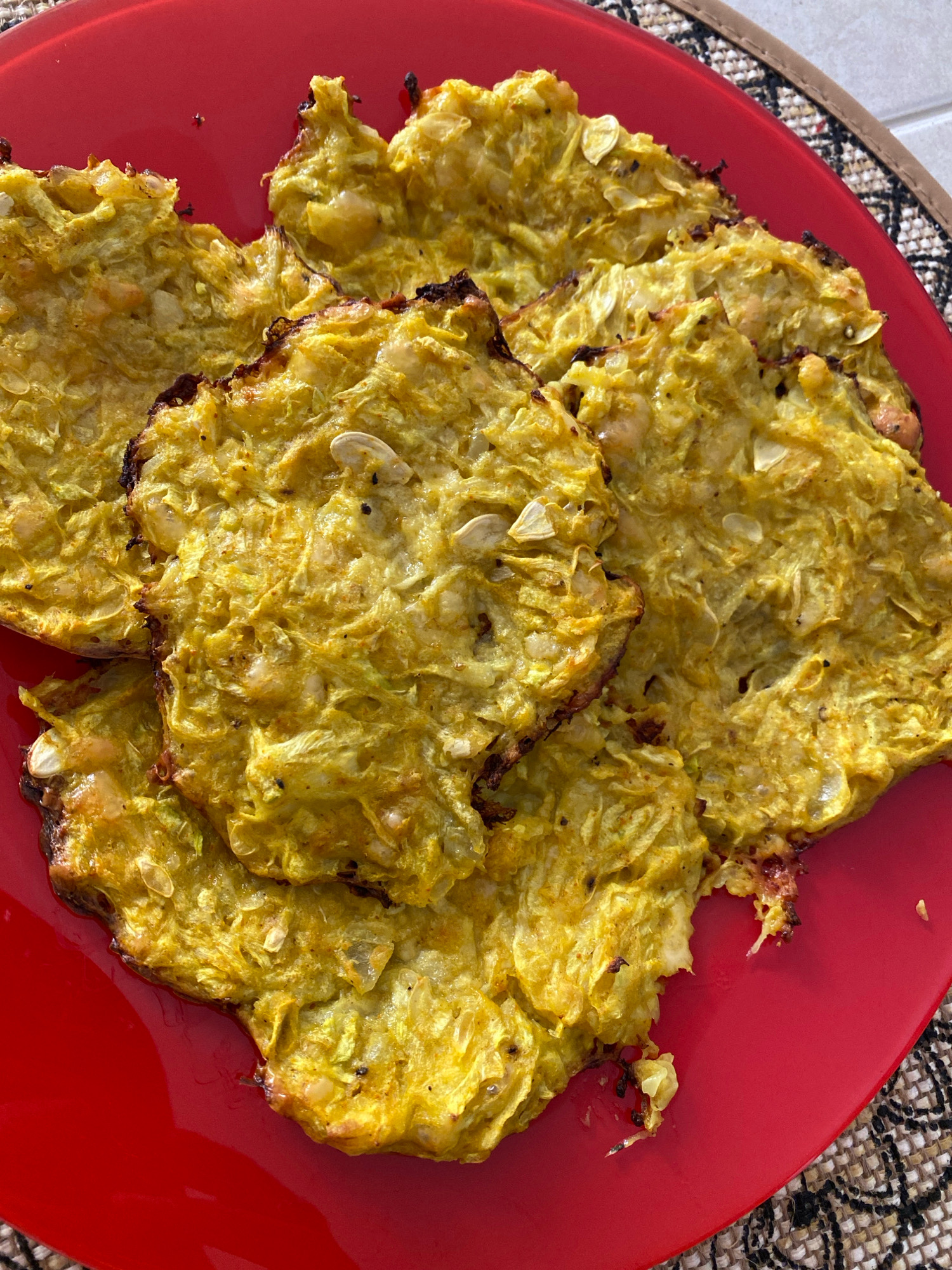 The image shows a close-up, overhead view of several golden-yellow, flat, savory pancakes or fritters piled on a vibrant red plate. The pancakes appear to be made from grated vegetables, possibly zucchini or squash, mixed with spices and perhaps some grains or seeds, which are visible as lighter flecks throughout the mixture. Some edges of the pancakes are darker, indicating they have been browned or slightly charred during cooking. The plate is resting on a woven placemat with a geometric pattern. There are no people or specific environmental details that would indicate a location in Lida, Belarus.