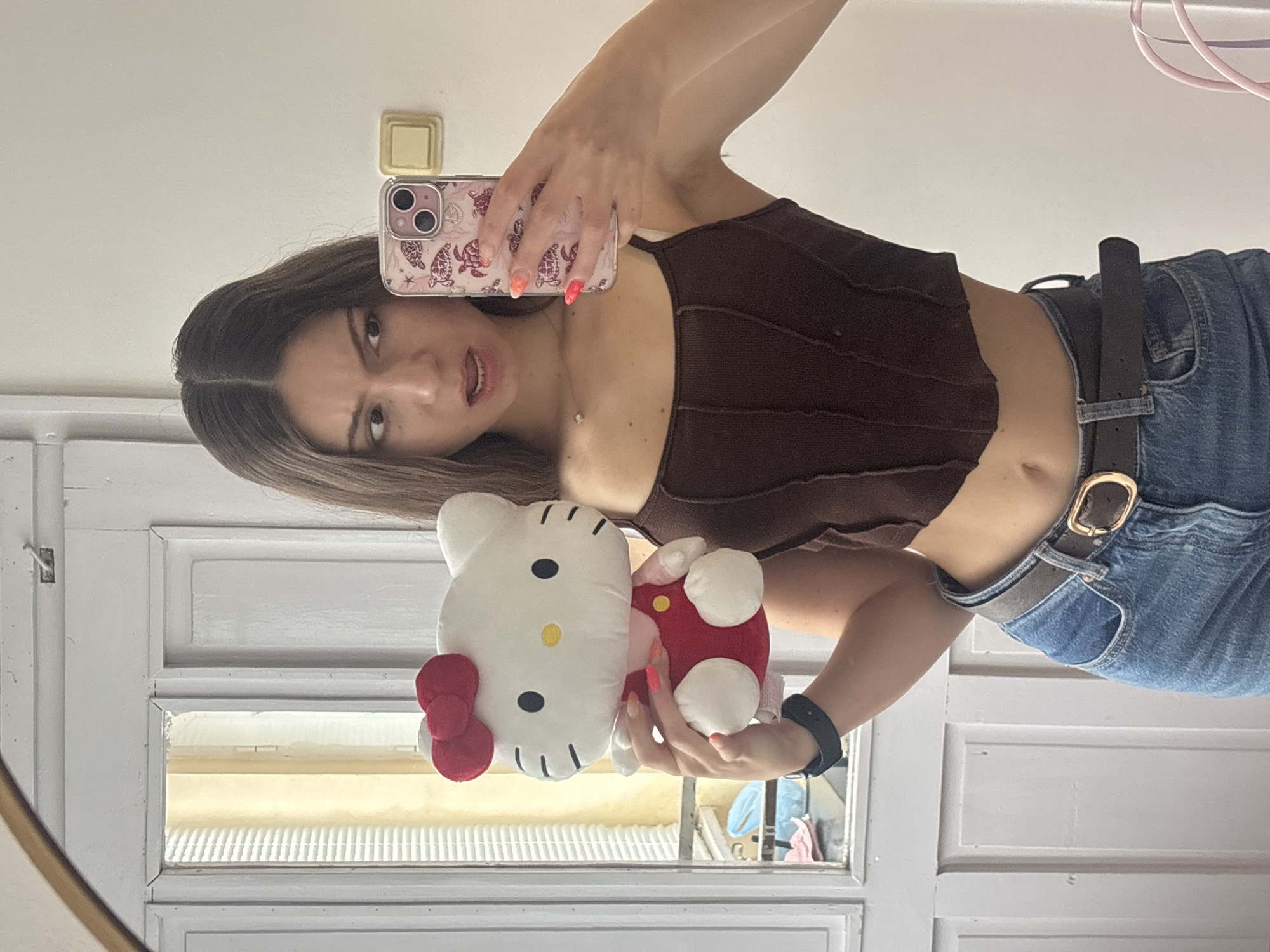 A young woman is holding a Hello Kitty plush toy while taking a selfie in front of a mirror. She is wearing a brown top, jeans with a brown belt, and a watch. The phone case is pink with a turtle pattern. The location appears to be indoors, possibly a room in a residence. There is a white door with window panels visible in the background. There is no verifiable evidence to determine that the location is in the city of Kaspichan, Bulgaria.