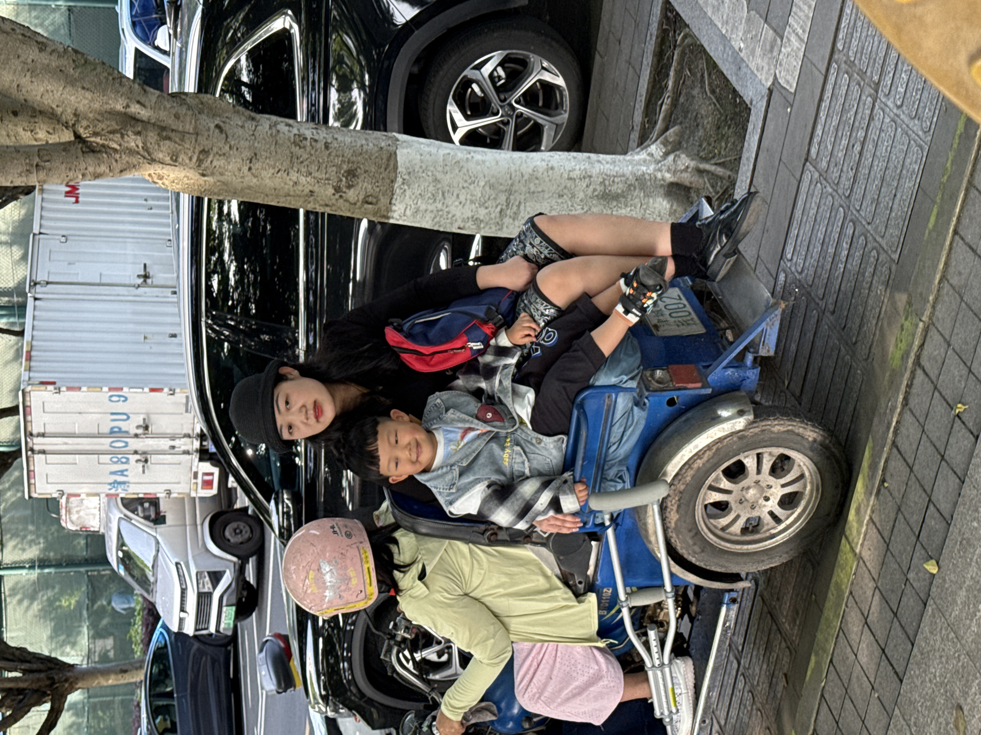This image captures an outdoor scene, likely on a street or sidewalk in Jiangjin, China. The primary subjects are an adult woman and two children interacting with a blue, three-wheeled motorized vehicle.
An adult woman with long dark hair, wearing a black beanie and a black long-sleeved top, is resting against the side of the blue vehicle. A red fanny pack is slung over her shoulder, and she looks directly at the camera with a calm expression. Next to her, a young boy with dark hair, wearing a denim jacket over a plaid shirt, sits smiling in the front passenger area of the blue vehicle, leaning against the woman. Partially visible in the driver's position is another person, possibly a child, wearing a light green top and pink shorts or skirt. A pink helmet with "JHL" written on it rests nearby.
The blue motorized vehicle has a single front wheel and two rear wheels, resembling an adapted scooter or tricycle, and a crutch is attached to its side. Visible text on the vehicle includes "200R" and "B1010Z".
In the background, a black SUV is parked, with its wheel visible. Further back, a white delivery truck is parked, displaying the text "JM" and "XA80PU91". A prominent tree trunk extends across the middle ground. The scene is set on paved ground, featuring tactile paving for pedestrians.
The activity appears to be a leisurely pause or waiting moment during the daytime under clear weather. The expressions of the woman and child convey a relaxed atmosphere.