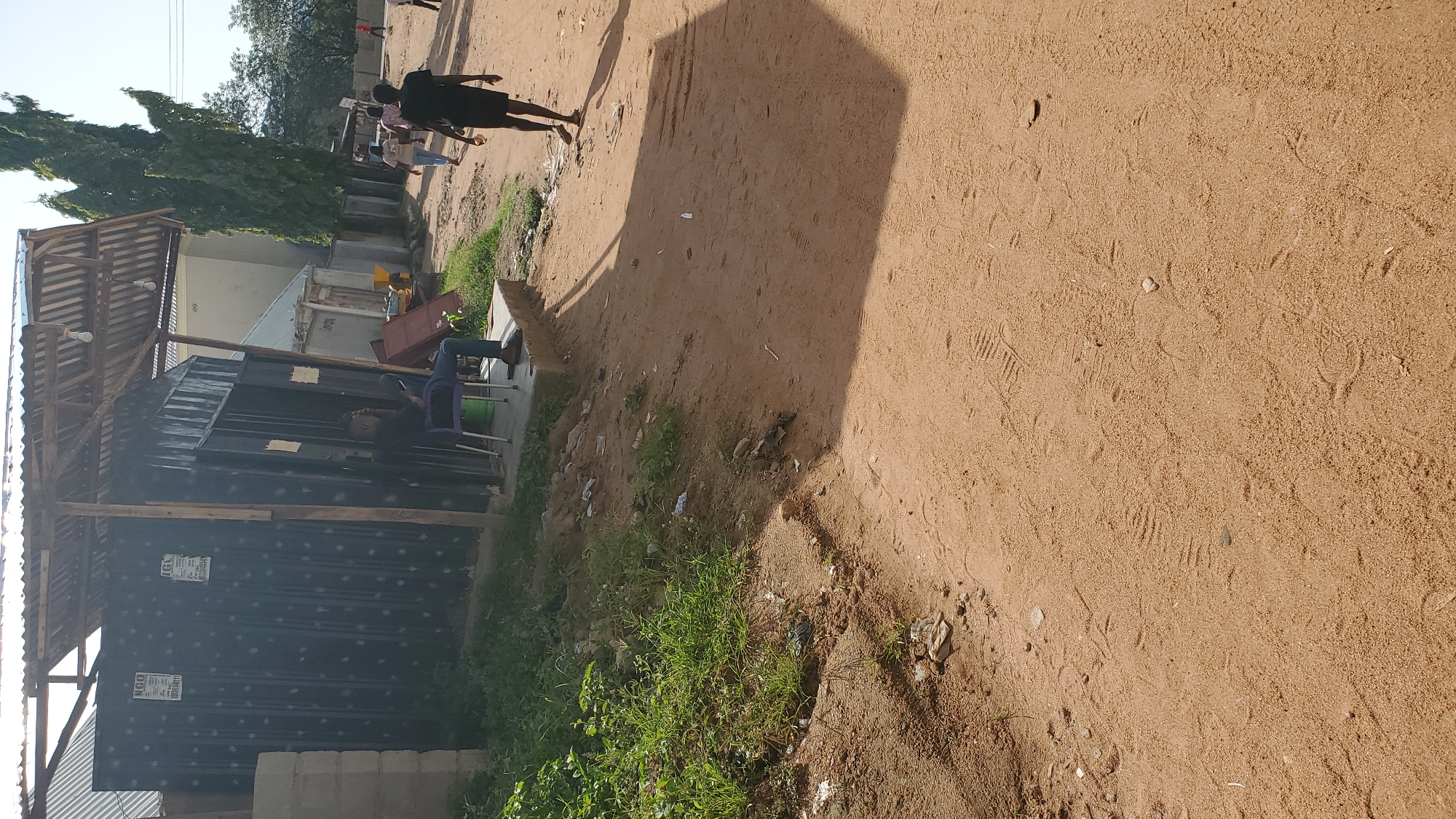 A man is sitting on a purple plastic chair outside a black corrugated metal structure in Jalingo, Nigeria. The structure appears to be a kiosk or shop, with a corrugated metal roof supported by wooden poles. The man is wearing a black t-shirt and blue jeans, and he is looking down at his phone. The ground is sandy and there are a few scattered pieces of trash. In the background, there are trees and other buildings. Further down the sandy path, another person, dressed in a black t-shirt and black shorts, is walking away from the camera. Several other people are visible in the distance, walking along the path. The overall scene depicts a typical street scene in a Nigerian city.