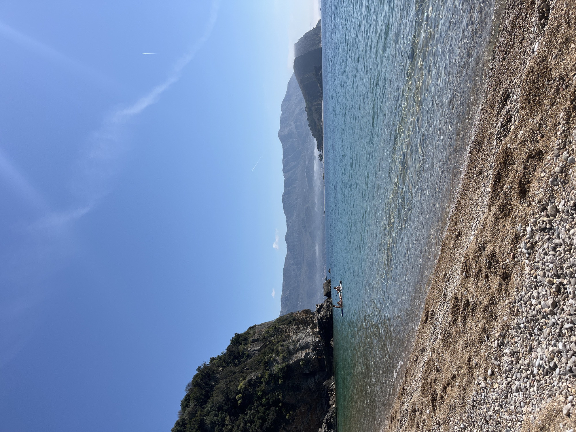 The image shows a coastal scene. The sky is blue, with a contrail visible. A mountain range is present in the background. A rocky outcrop covered in vegetation is on the left. The shoreline consists of a pebble beach where the sea gently meets the land. There are two individuals on a paddleboard near the rocky outcrop. The location may be near Budva, Montenegro, given the landscape and coastal features.