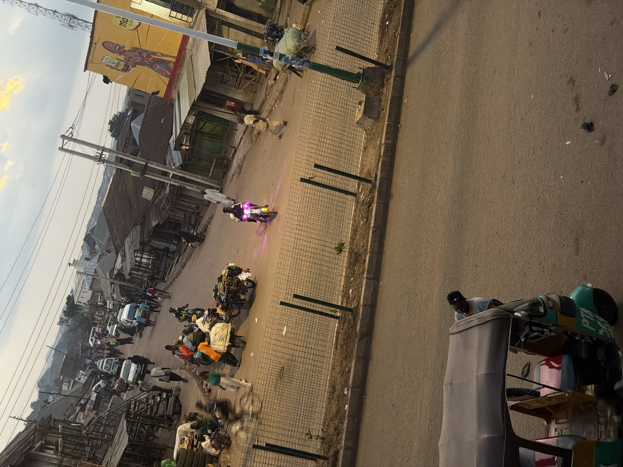 The image depicts a street scene in Bukuru, Nigeria, likely a marketplace or commercial area. The time of day appears to be late afternoon or early evening, given the lighting. In the foreground, a three-wheeled auto-rickshaw, also known as a Keke Napep, is parked. A man wearing a light blue t-shirt and a black baseball cap is leaning over its hood, appearing to be working on it or attending to it. The auto-rickshaw is predominantly green with yellow accents and displays the license plate "PTA 269".
The middle ground shows a busy street with various people and vehicles. A motorcycle with bright pink lights is being ridden down the street. Several individuals are walking, and some are pushing carts laden with goods, suggesting commercial activity. Among the goods are textiles and other merchandise. A cart piled high with what appears to be clothing or fabrics is visible, as are several other carts with various items. In the background, there are numerous buildings with corrugated metal roofs, typical of a market environment. A large mural advertising "Ma" is painted on the side of a yellow building on the right. The street is lined with stalls and shops, some of which appear to be open for business. A chain-link fence runs along the side of the street, separating the road from a raised area with some sparse vegetation. Further in the distance, hills and more buildings can be seen. Power lines crisscross the sky, and a cell tower is visible in the upper right portion of the image.