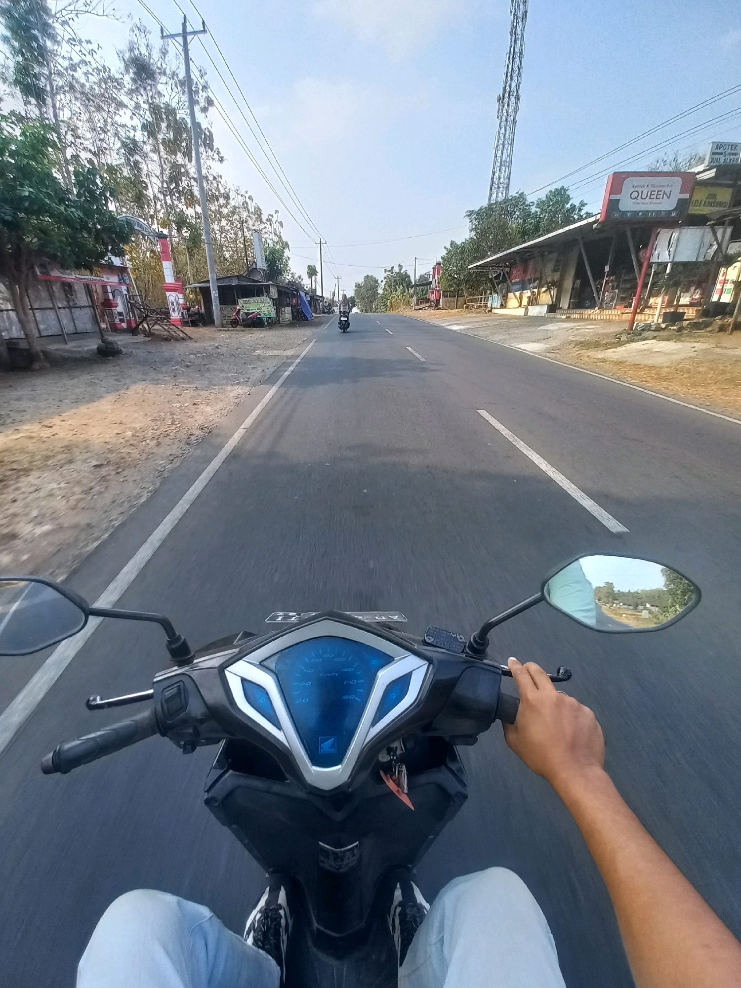 The image shows a first-person perspective of a person riding a scooter on a paved road in Baturetno, Indonesia. The road has a white line marking its center and right edge. On the left side of the road, there is a dusty area with some trees and small buildings, including one with a red and white striped structure. Further down the road on the right, there are several shops with awnings, one of which has a sign that reads "QUEEN". A tall telecommunications tower is visible in the background. The scooter has a blue digital dashboard and a hand is seen on the right handlebar. The rider is wearing light-colored pants and dark shoes. The sky is clear and blue, indicating daytime.