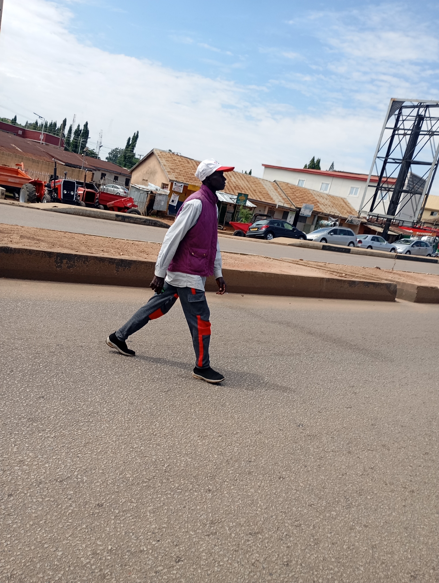 A full-shot photograph captures a man in a purple vest, white long-sleeved shirt, and dark pants with orange accents walking on an asphalt road. He wears a white baseball cap. In the background, several tractors are parked on a dirt patch adjacent to the road, and a billboard structure looms. The environment suggests an urban setting with buildings and parked cars further in the distance, under a partly cloudy sky. The scene is devoid of any overt signs indicating the specific location as Gura Top, Nigeria.