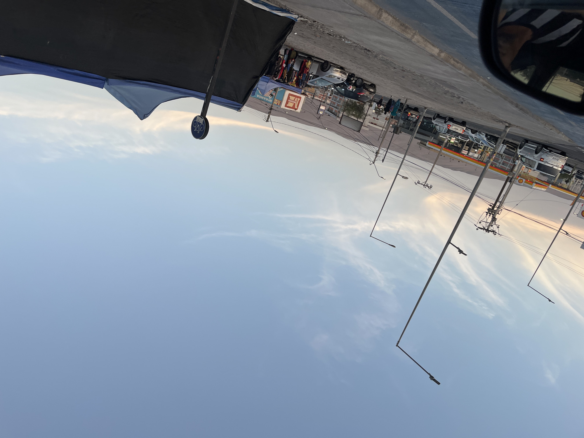 The image depicts a street scene in Arica, Chile. The view is from inside a vehicle, with a portion of the side mirror visible in the upper right corner.
On the left side of the frame, a dark blue and black canopy is partially visible. To the right of the canopy, there appears to be a street market with vendors selling goods, including clothing.
Further back, a gas station is visible, identified by the Shell logo. Cars are parked at the gas station, and other vehicles are moving along the road. Power lines stretch across the sky. The sky is mostly clear with some scattered clouds.