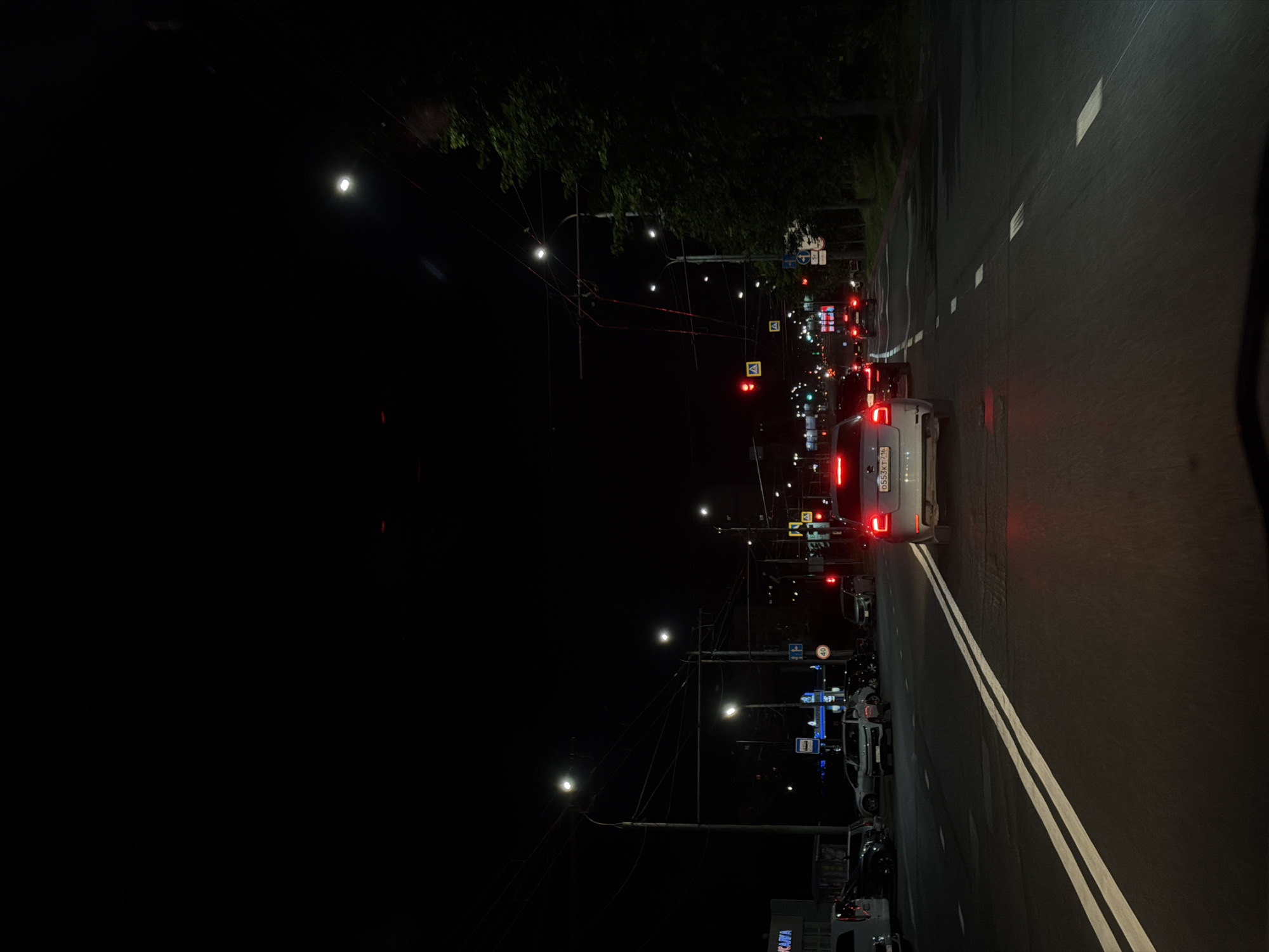 A nighttime, slightly elevated view of a street in an urban setting. The road is paved and marked with double white lines, indicating multiple lanes. Streetlights illuminate the scene, casting a glow on the asphalt and surrounding trees. Several cars are visible, both parked on the side of the road and in traffic. One car, a white sedan, is prominently in the foreground, its brake lights illuminated. Traffic lights are visible at an intersection, with some showing red. A pedestrian crossing sign is also present. Buildings line one side of the street, with a sign for "КАПЛЯ" visible on one. The overall atmosphere is one of a typical city street at night, with active traffic.