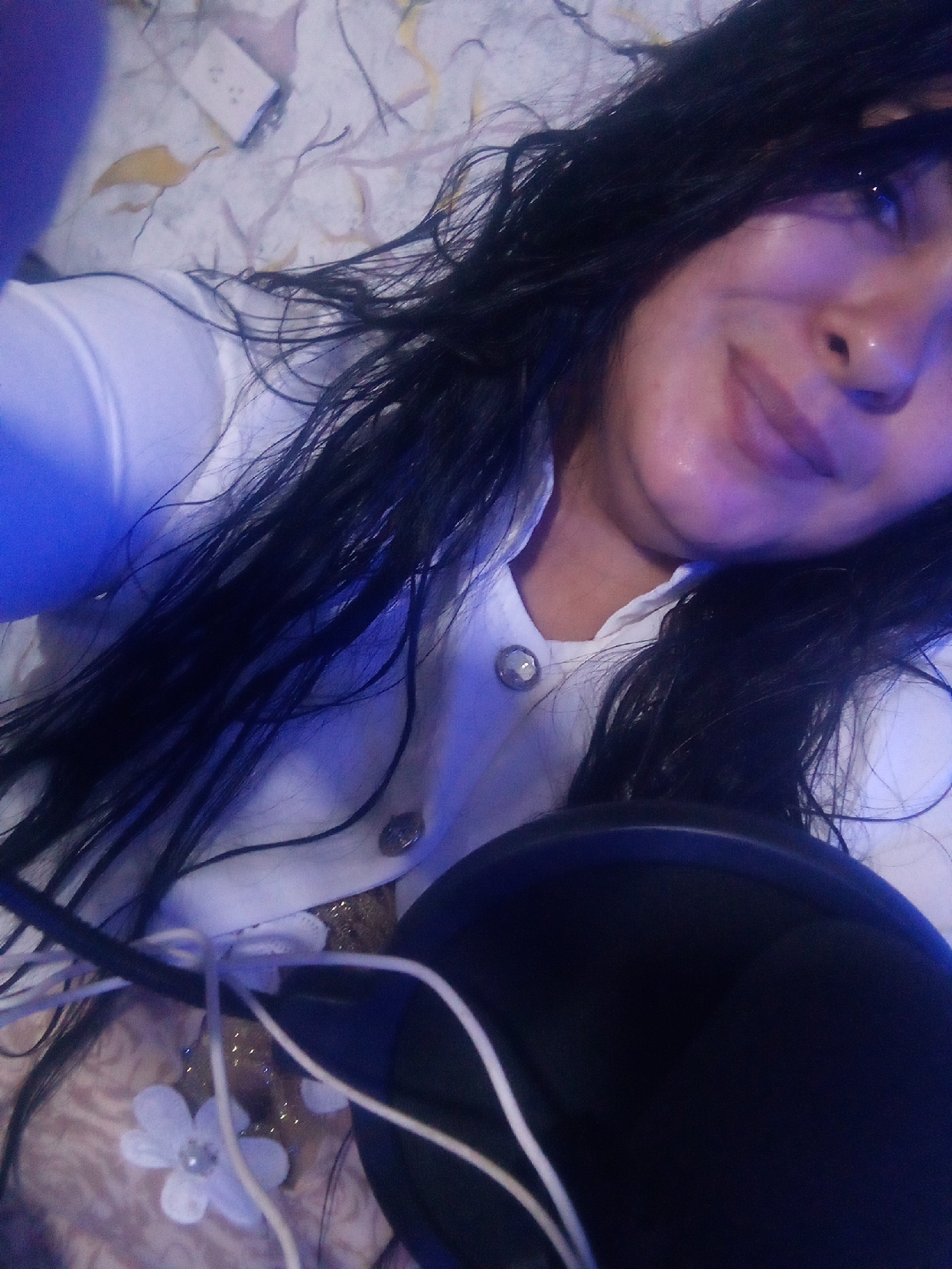 A selfie from a low angle shows a woman with dark hair and light skin. She is wearing a white button-down shirt. A large, round, black object with white wires extends from the bottom of the frame, partially obscuring the scene. A white flower with a pearl center is near the black object and wires. The background consists of a patterned surface in shades of beige, white, and yellow. In the upper-left corner is a white electrical outlet.