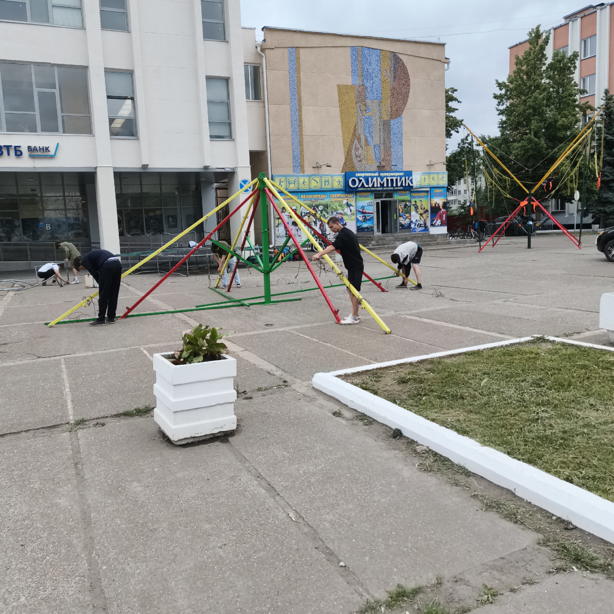 In Chaikovsky, Russia, on a cloudy day, a group of individuals is in the process of setting up an outdoor recreational structure, likely a bungee trampoline, on a paved plaza in front of a commercial building. The building has a mosaic mural on its facade and signage for "ОЛИМПИК" (Olympic) sports supermaket. Several people, dressed in casual clothing, are actively engaged in assembling the equipment. One person, wearing a black shirt and shorts, is adjusting the yellow elastic cords of the trampoline. Another, in a white shirt and dark shorts, is bending down and appears to be connecting or securing parts of the structure. A third person, in a green shirt, is also working on the base of the setup. In the foreground, a white planter box with a green plant sits on the pavement. The overall scene depicts an effort to install an amusement attraction, possibly for a public event or a business.