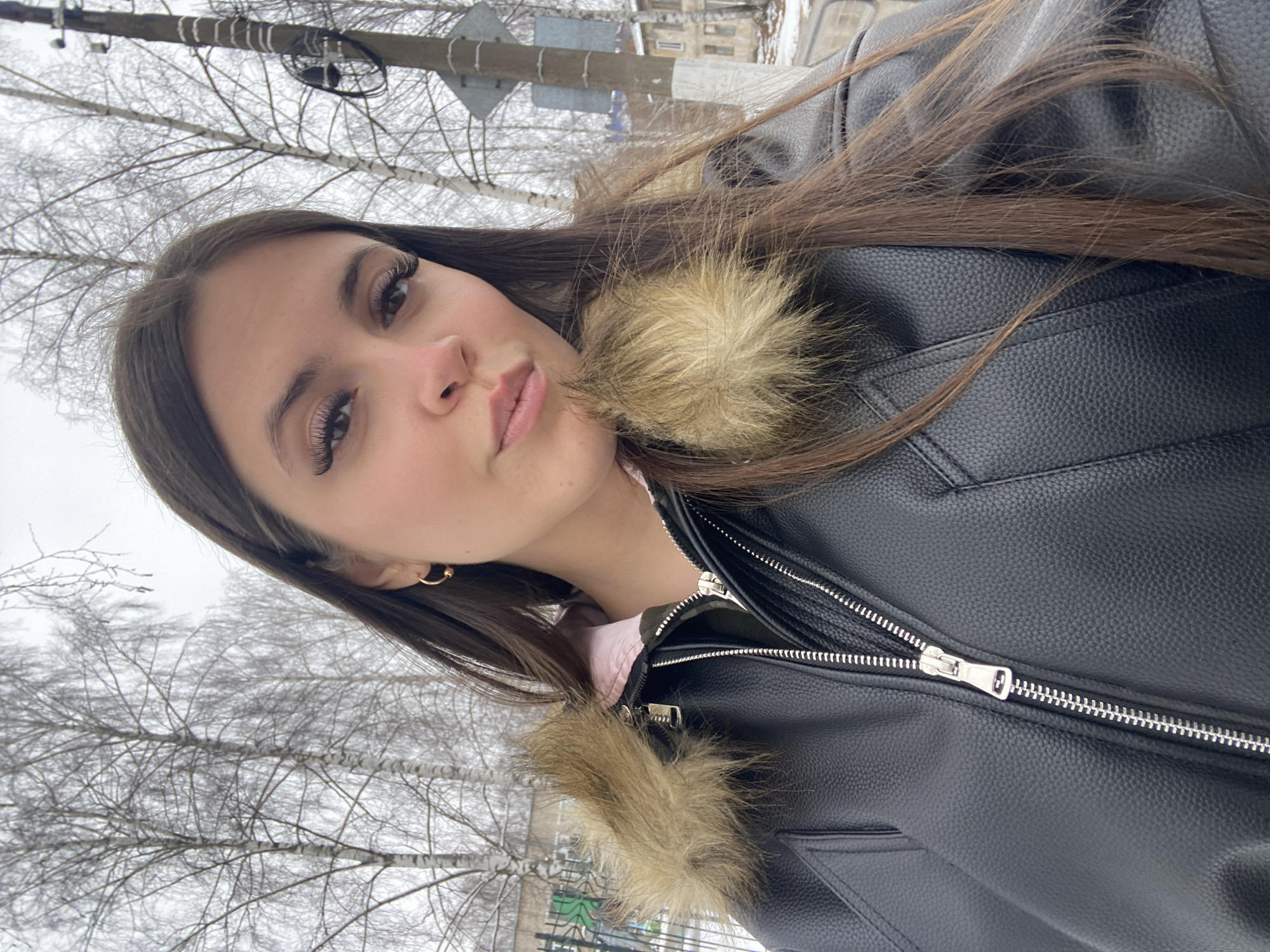 The image is a selfie of a woman wearing a black leather jacket with fur trim, taken outdoors. The background reveals bare trees, possibly indicating winter or early spring. Utility poles and buildings suggest an urban environment. Based on the metadata, the image was taken in the city of Chaikovsky, Russia.