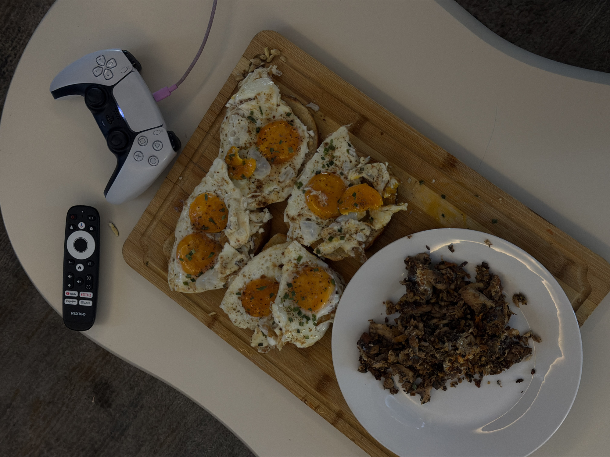 An overhead shot shows a table with a meal and a video game controller in London, United Kingdom. The table surface is light-colored and slightly curved. In the upper left corner, a black TV remote control with red buttons sits on the table. To its right, a white PlayStation 5 controller is plugged in with a purple cable. The main focus is a wooden cutting board with four pieces of toast topped with fried eggs and chives. To the left of the cutting board, a white plate holds a pile of what appears to be shredded, seasoned meat.