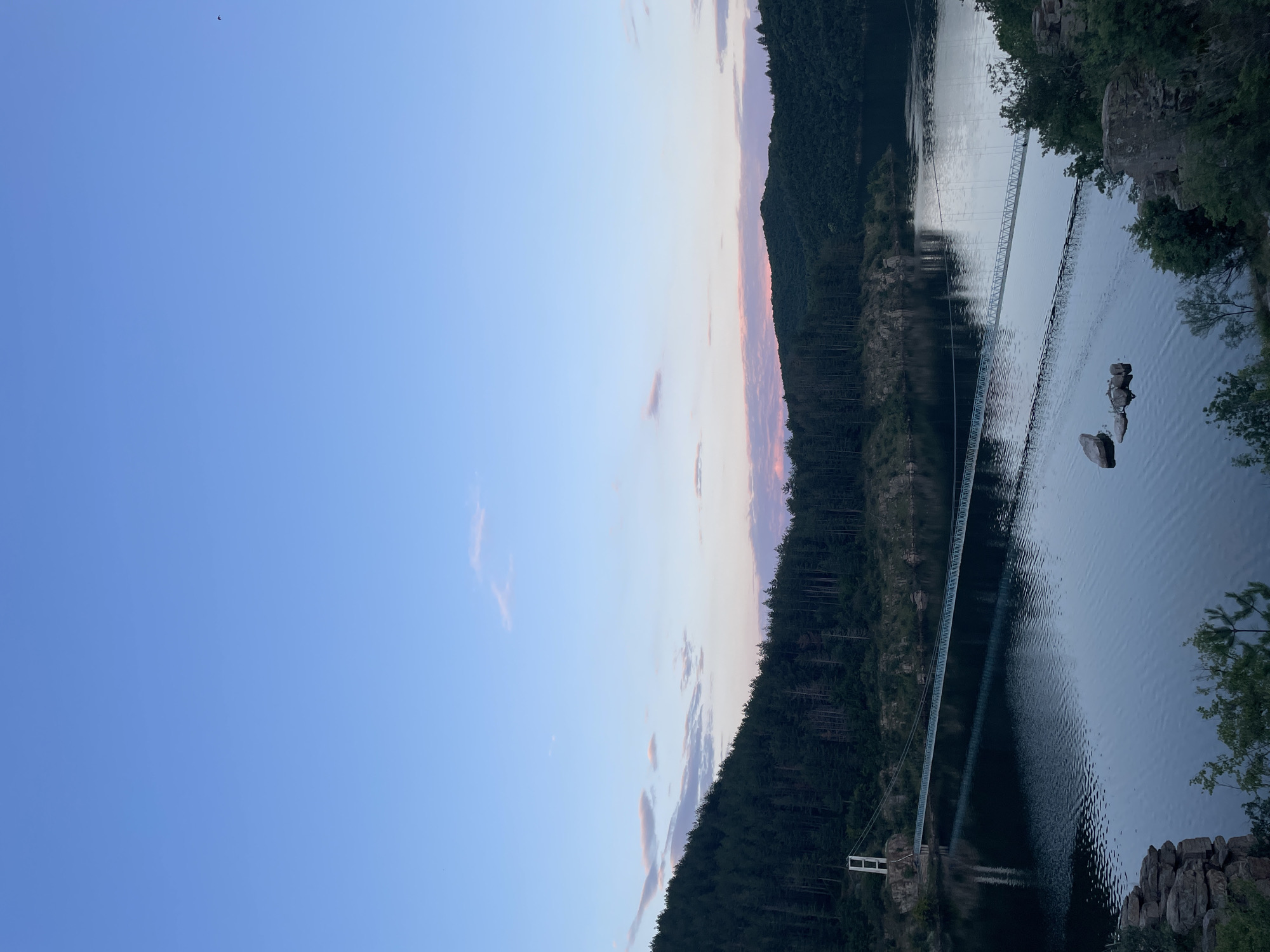 This image depicts a wide shot of a suspension bridge spanning a tranquil body of water, likely a reservoir or lake, surrounded by lush, dark green pine forests that climb the hillsides. The sky above is a soft gradient of blue, transitioning to lighter hues with streaks of pink and lavender clouds, indicating either dawn or dusk. The bridge, with its white towers and metallic walkway, is the dominant man-made structure, its reflection visible on the still surface of the water. A few scattered rocks break the monotony of the water's surface. The scene conveys a sense of serene natural beauty, characteristic of the area around Suhovo, Bulgaria, known for its scenic landscapes and reservoirs.