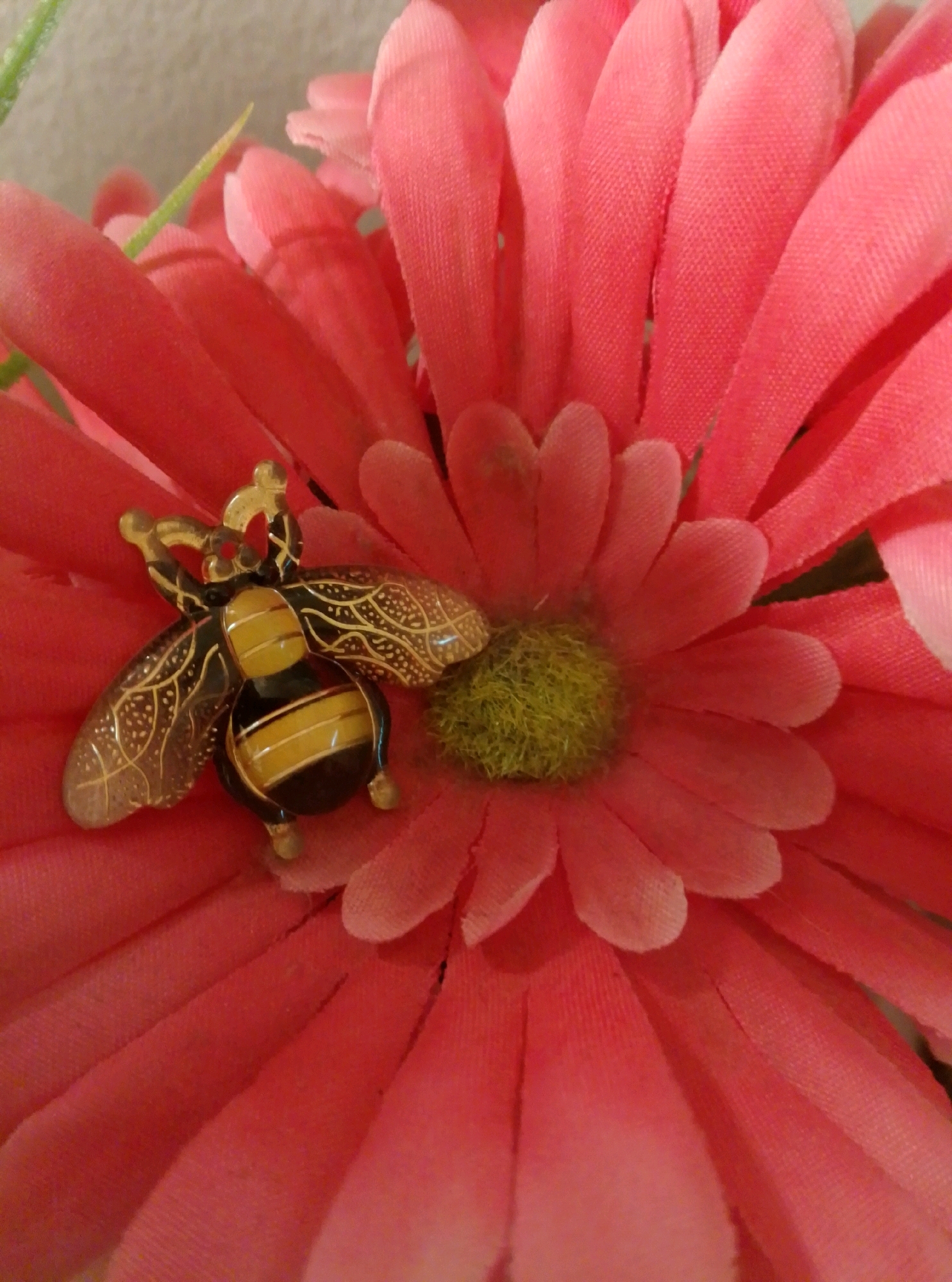 A close-up, eye-level shot focuses on a decorative bee pin resting on a pink, artificial flower. The flower, likely a gerbera daisy, has numerous soft, fabric petals that are a vibrant pink hue. The bee pin is detailed with black and yellow stripes on its abdomen, delicate gold wings with intricate etched patterns, and a red gem for its eye. The flower's center is a fuzzy, light green pistil. The background is a neutral, textured off-white wall. There are no people, actions, or interactions depicted. The scene is static and appears to be a still-life arrangement. The location in Wedel, Germany, is not discernible from the visual information provided in the image.