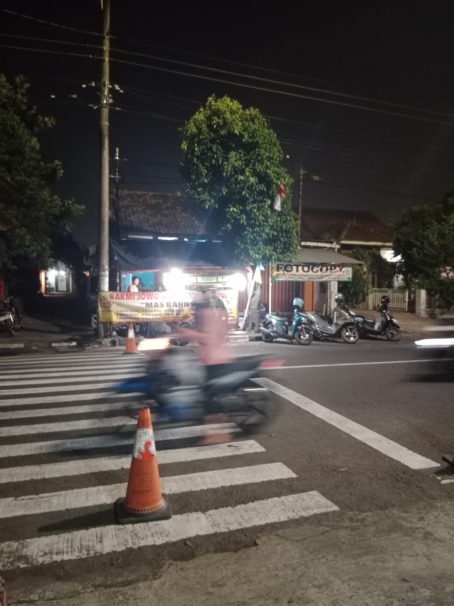 This is a medium-shot, eye-level, slightly blurry night photograph taken at a crosswalk in the city of Yogyakarta, Indonesia. In the foreground, an orange traffic cone stands on the zebra stripes of the crosswalk. To the left of the cone, a person on a motorcycle is blurred as they move across the crosswalk. In the background, a street is lined with several parked motorcycles and a food stall with a banner that reads "BAKMI JOWO MAS RAHNO". Above the food stall, a "FOTOCOPY" sign is visible. Trees and utility poles with numerous wires are scattered throughout the scene. The lighting is dim, with artificial lights illuminating the food stall and parts of the street.