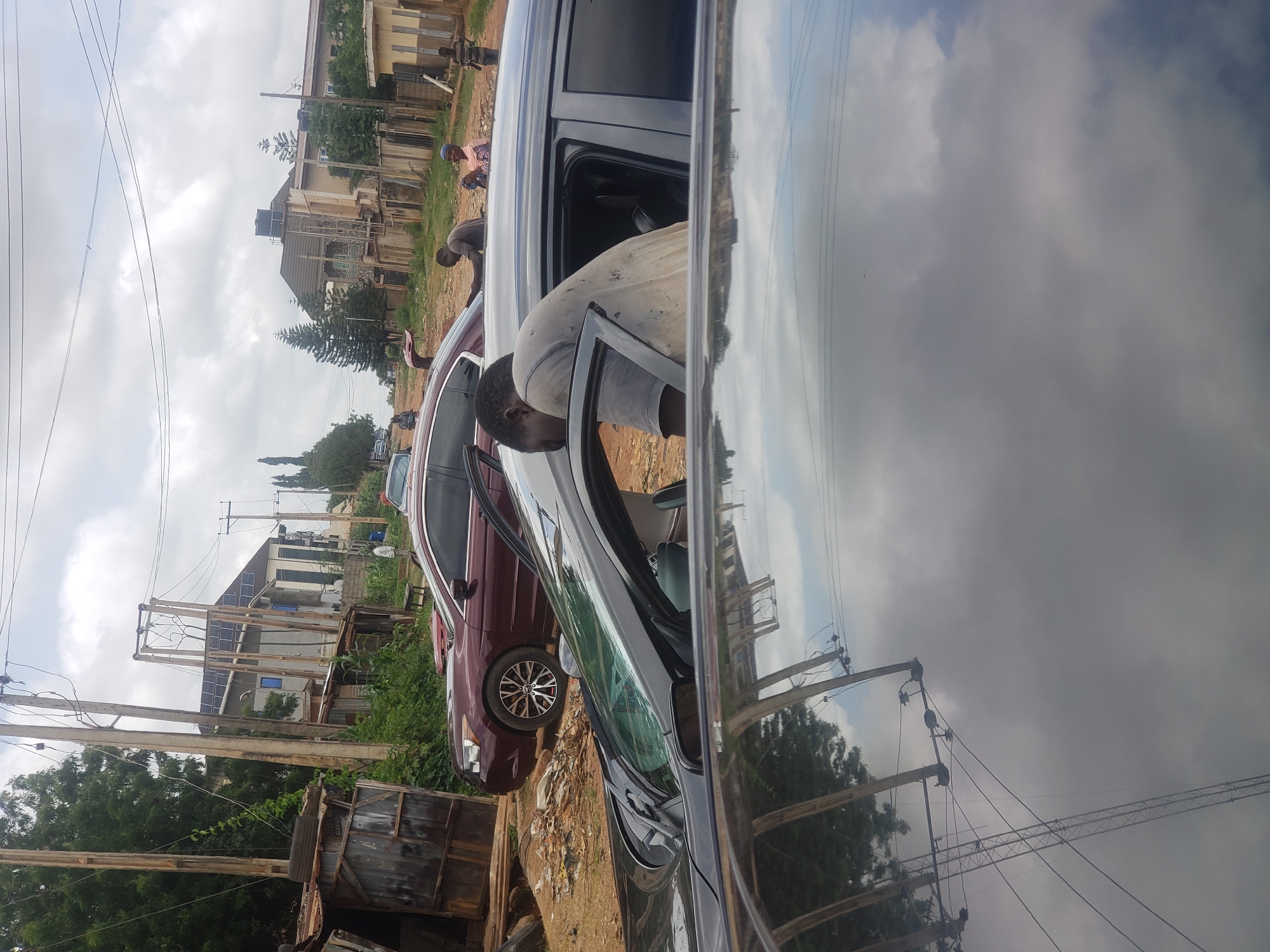 In Narayi, Nigeria, a man in a light-colored, stained shirt is leaning into the open driver's side of a dark-colored sedan. The car's interior is visible, with dark seats. The man appears to be working on or interacting with the car's interior. In the background, a burgundy sedan is parked. Further in the distance, several buildings with solar panels on their roofs are visible, along with telephone poles and lush green trees. A dirt road is present in the foreground, and debris is scattered around. Several other people are present in the background, some standing and observing. The sky is overcast with clouds.