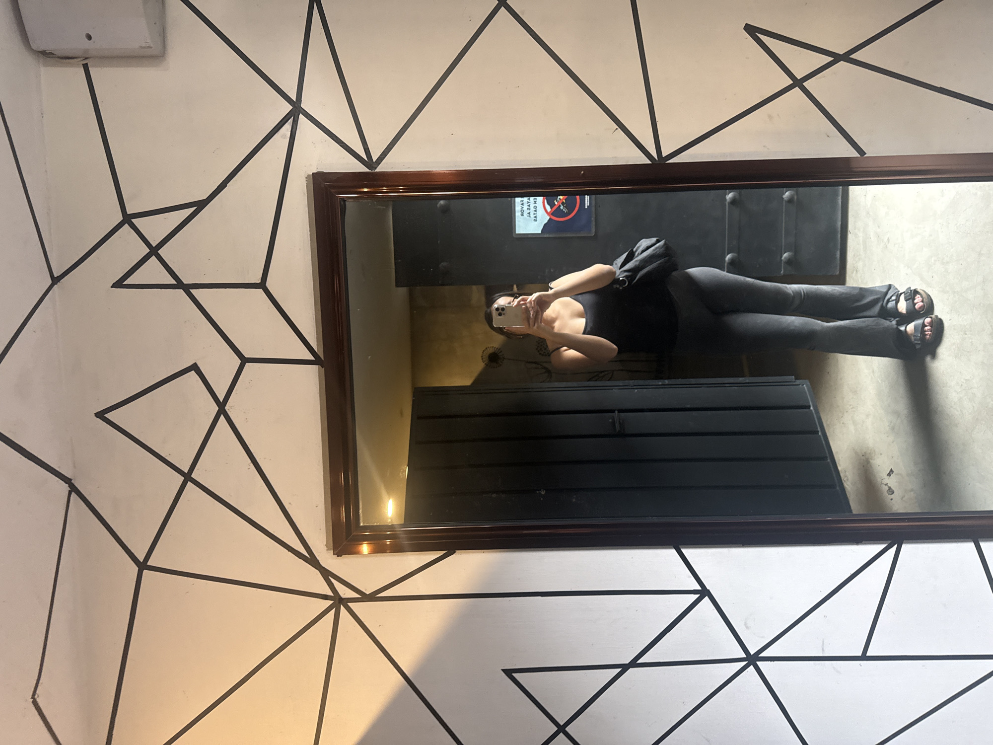 A person is visible in the reflection of a rectangular mirror with a brown frame that is attached to a wall with a geometric black and white design. The person is holding a smartphone and appears to be taking a photograph or video of themselves in the mirror. The individual is wearing a dark-colored top and pants, and a dark-colored shoulder bag. The background reflected in the mirror shows a room with dark, horizontal slat walls, a metal door with rivets, and a concrete floor. A sign with a no smoking symbol is visible near the top of the reflection. The location is San Andrés Cholula, Mexico.