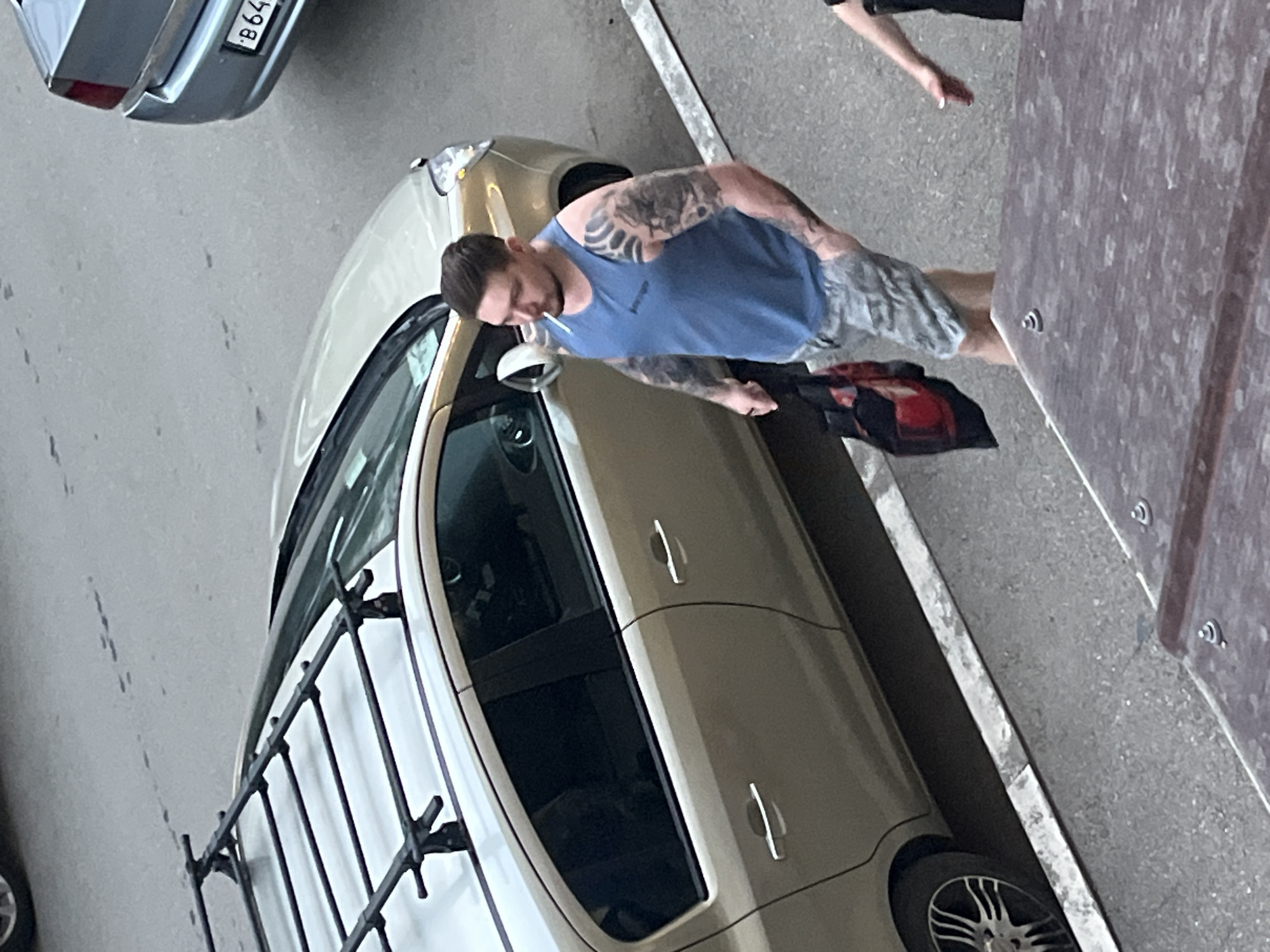 A man with tattoos on his arms and neck is walking next to a tan sedan. He is wearing a blue tank top and grey camouflage shorts. He is holding a red and black bag in his left hand and a cigarette in his mouth. The car has a roof rack. Another car, a silver sedan, is parked behind the tan car. The scene appears to be on a paved street in Krasnodar, Russia, with a raised sidewalk or curb visible. The foreground shows the edge of a wooden structure, possibly a table or bench.
