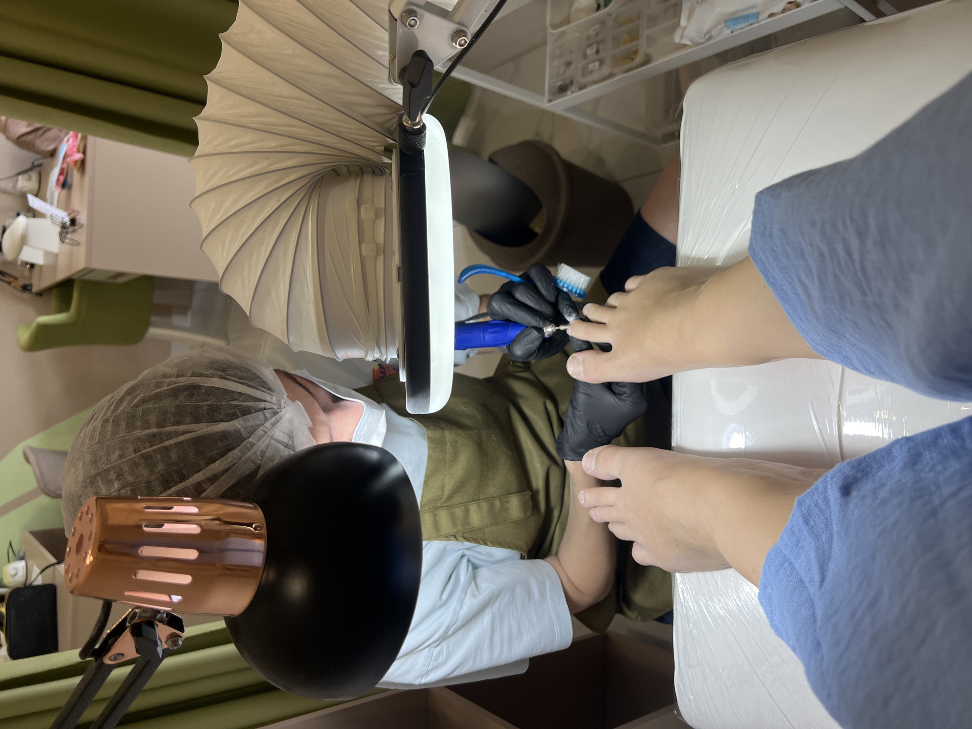 A person wearing a hairnet and face mask is providing a pedicure in a room. A drill and brush are used on the patient's feet, which are resting on a white, wrapped cushion. The person providing the pedicure is wearing black gloves. An extraction device hangs above the area being treated. In the room are visible a black and copper lamp, drawers, and a green chair. Based on visual clues, the location is likely a salon or spa. The city of Podgorica, Montenegro cannot be verified from this image alone.