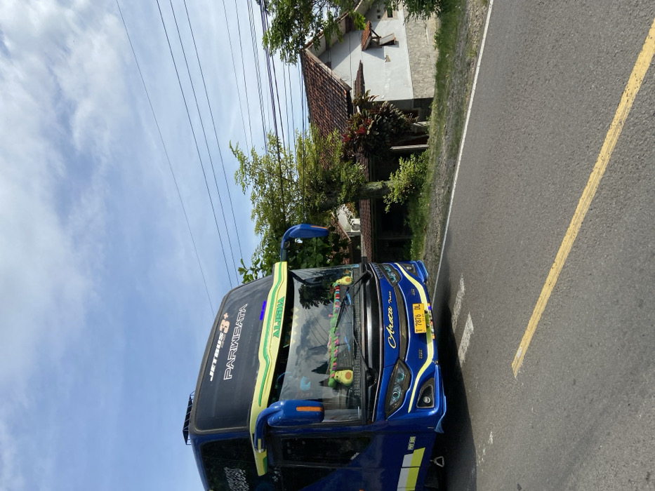 The image shows a blue and yellow passenger bus labeled "Jetbus 3+ Pariwisata" and "Alica" traveling on a paved road with yellow lines. The bus is driving away from the camera and is slightly angled to the right. The license plate reads "I 7876 DL." The bus is on a road next to a residential area with small, white houses with red tile roofs. The sky is blue with white clouds. There is a small, green bush in the foreground on the right. The location of the image is Sukatani, Indonesia, located at coordinates -6.635 latitude and 107.404 longitude. There are no people visible in the image. The image was likely taken during the day.