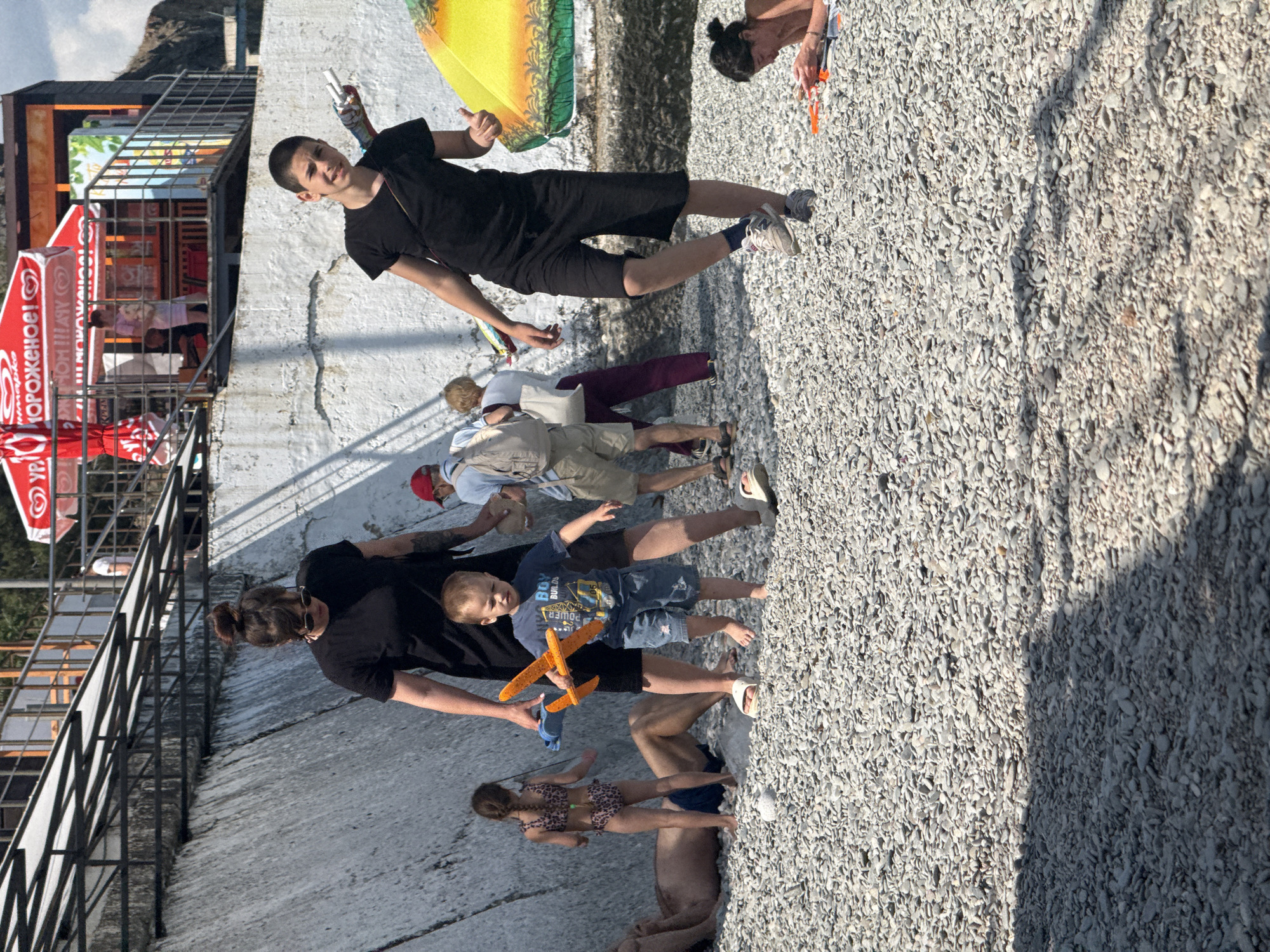 The image shows a sunny day at a pebble beach. In the foreground, a young boy in a blue shirt and denim shorts holds an orange toy airplane. To his left, a woman with dark hair in a bun, wearing a black t-shirt, stands next to him. Behind them, a young girl in a leopard-print swimsuit is partially visible. Further back, a man in a red cap and a backpack walks away from the camera, accompanied by another woman. To the right, a teenager in a black t-shirt and black shorts walks towards the camera, holding a white object. Another person is lying on a towel in the lower right corner of the image, with an orange object on the towel. A yellow and orange umbrella is visible on the right side of the frame. The background consists of a concrete wall with a waterfall-like structure, and structures that appear to be part of a beachside establishment with red umbrellas. The ground is covered with small pebbles.