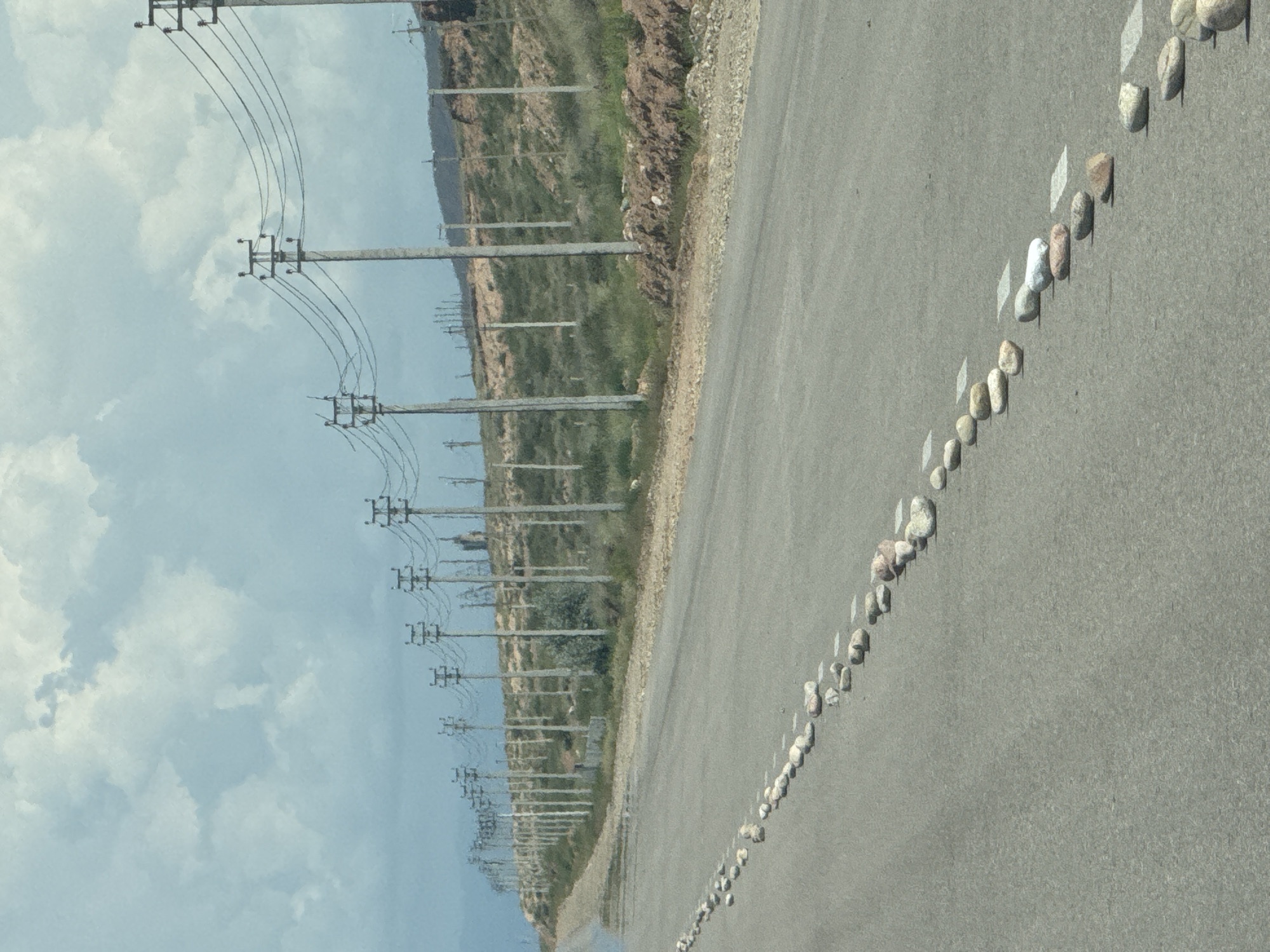 The image depicts a paved road in Kaji-Say, Kyrgyzstan, with a line of rocks placed along the white dashed lane marker on the right side of the road, extending into the distance. Overhead, a series of utility poles with wires stretches along a sloping embankment on the right side of the road, which is covered with sparse vegetation and some exposed soil. The sky above is partly cloudy. The scene is devoid of any people or vehicles.