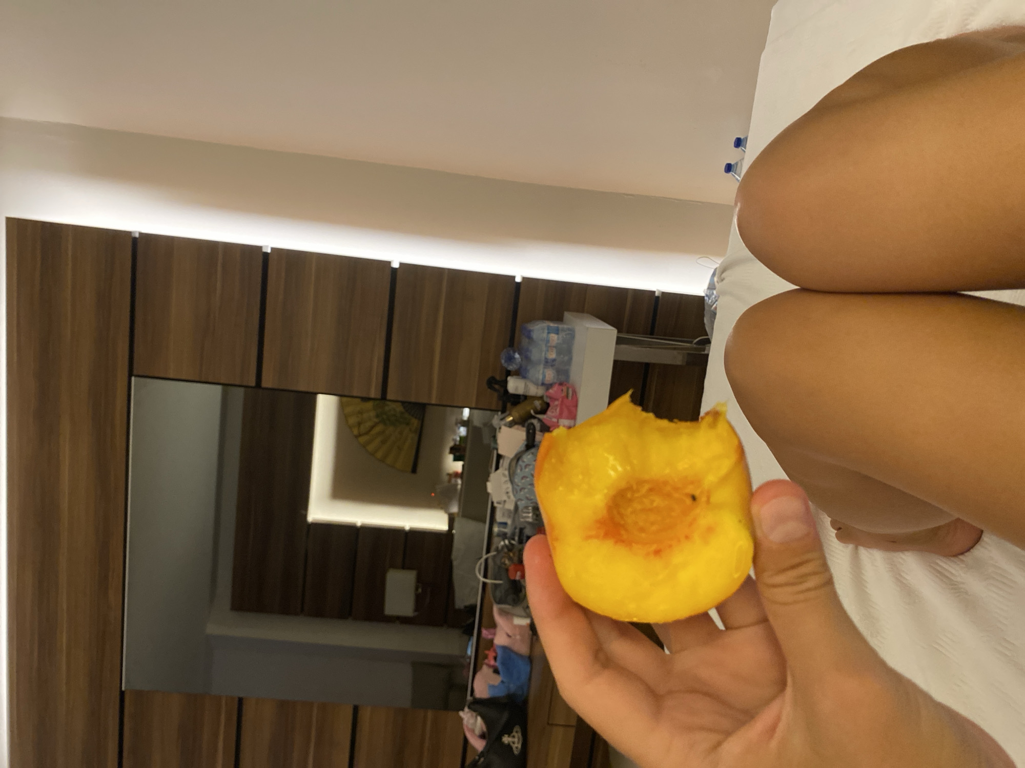 In the city of Aksu, Turkey, a person is holding a partially eaten peach over a bed. The person's legs are visible in the foreground, resting on white sheets. In the background, a dresser with a large mirror reflects a room. Items are scattered on the dresser, including bags, bottles of water, and a decorative fan. A bright light emanates from a fixture on the wall to the right.