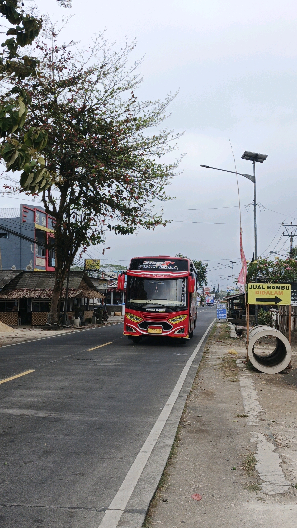 A red tour bus with the words "PUTRA KARYA" and "PARIWISATA" on its exterior drives down a two-lane asphalt road in Cibenda, Indonesia. The bus, model Voyager, displays a license plate "Z 7535 TH." On the left side of the road, a tree with sparse red and green leaves overhangs a row of small buildings with thatched roofs, one of which displays signage that is partially visible. A modern building with gray and red facade is visible behind these structures. On the right side of the road, a bright yellow sign reading "JUAL BAMBU DIDALAM" with an arrow pointing to the right is mounted on a wooden post. Next to the sign are two large concrete culverts. A solar-powered street lamp is also visible on this side of the road. The sky is overcast. The scene is captured from a slightly low angle, looking down the road as the bus approaches.