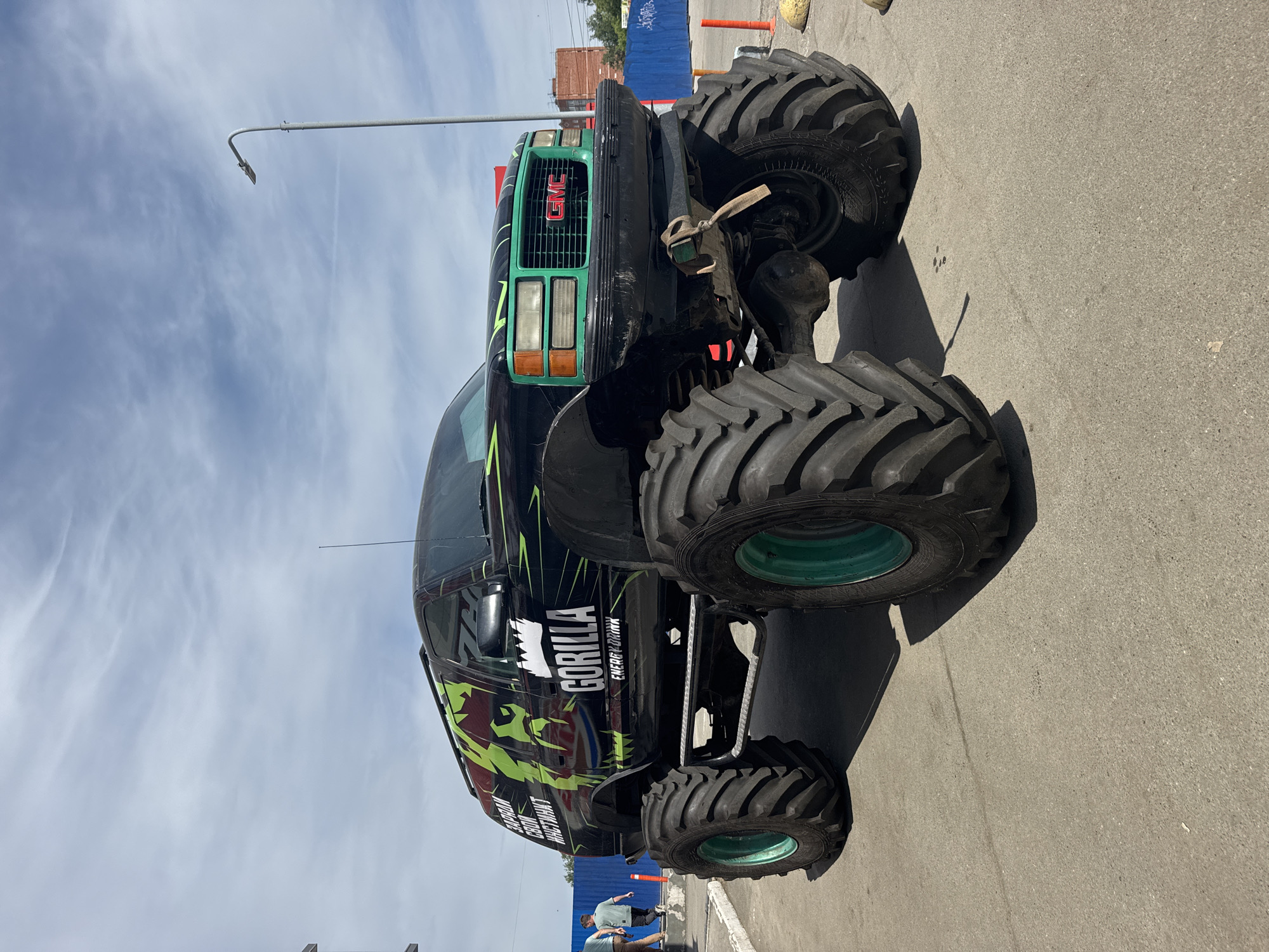 A monster truck branded with "Gorilla Energy Drink" is parked on an asphalt surface. The truck is black with green accents and large, heavy-duty tires with green rims. The front of the truck has a green grille with a red GMC logo. A light pole stands in the background. Two people are near the front of the truck, near blue and orange construction barriers. Based on the visible details, the location is consistent with an urban environment, possibly in Izhevsk, Russia.