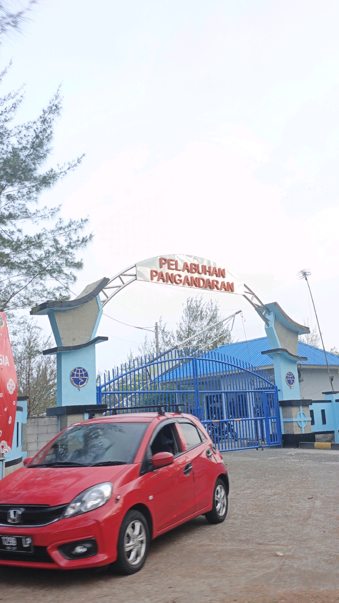 A red Honda Brio hatchback is parked in front of a blue gate at Pelabuhan Pangandaran, located in Desa Karangjaladri, Indonesia. The car has a roof rack. The gate is part of a larger structure that includes a blue building with a light blue roof and two blue pillars supporting an archway. The archway has a banner that reads "PELABUHAN PANGANDARAN" in red lettering. To the left of the gate, there is a red and white banner with text that is partially visible. Pine trees are present in the background. The sky is overcast.
