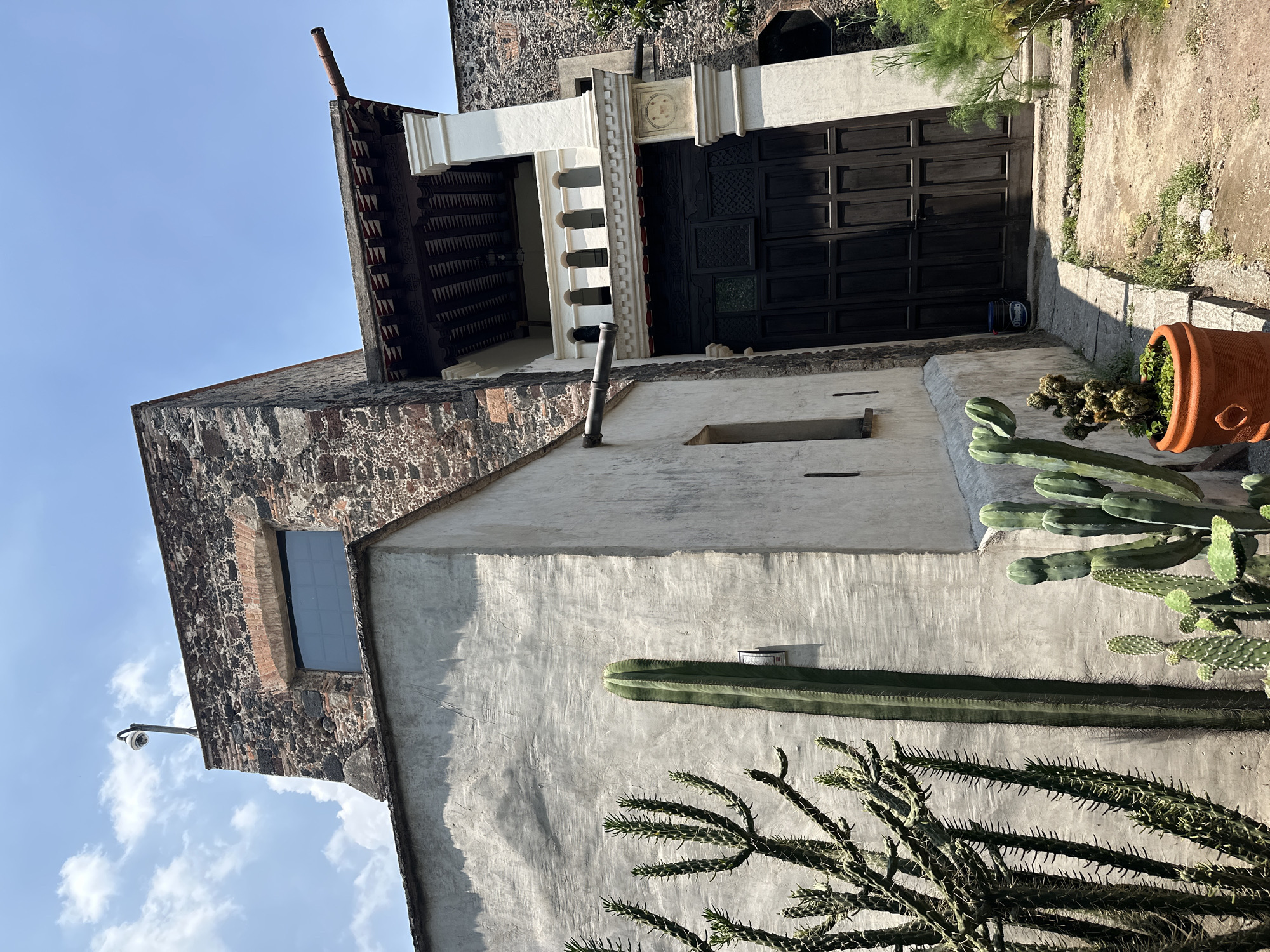 The image depicts the exterior of a building, likely a residence, showcasing a blend of traditional and rustic architectural elements, set against a clear blue sky.
The main subject is a two-story structure. The upper section features walls constructed from dark, rough-hewn stone, while the lower section is covered in a lighter, textured stucco or concrete. A dark wooden roof overhang, with a geometrically patterned underside, extends from the top of the building. Below this, a large, dark brown wooden double gate or garage door is prominent, featuring paneled sections. Above and to the side of the gate, decorative white architectural elements are visible, including a small balustrade and a larger, almost cross-shaped lintel or frame.
The foreground and mid-ground are rich with various cacti. A very tall, slender columnar cactus stands against the stucco wall. In the immediate foreground and to the lower right, a bushy, spiky cactus with numerous branches is visible. Another type of cactus, possibly an Opuntia or Cholla, is planted in a round, red terracotta pot near the gate.
Notable details include a small rectangular window with multiple panes set into the stone wall on the upper level, and a very narrow, vertical opening in the stucco wall below. A white security camera is mounted on the upper left corner of the stone wall. The ground in front of the gate appears to be a rough, unpaved or concrete path.
The time of day is bright, with sunlight casting distinct shadows, suggesting either late morning or afternoon. The sky is clear blue with a few scattered white clouds, indicating fair weather. The architectural style and prominent cacti suggest a location in a warm climate, consistent with Mexico City.
Visible text includes the word "PREMIER" on a blue bucket situated near the base of the wooden gate, and the word "ALARMA" on a small white sign affixed to the wall next to the gate.