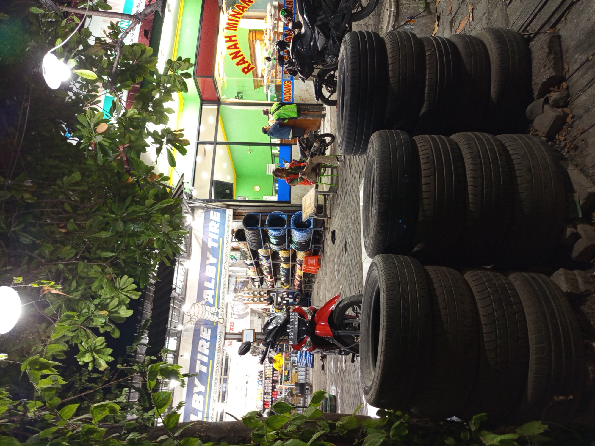 The image captures a nighttime scene outside a tire shop named "ALBY TIRE" and a food establishment. The food establishment, identified as "RANAH MINANG PADANG," displays food items behind a glass counter. In front of ALBY TIRE, several stacks of tires are prominently displayed, with a red motorcycle parked in the foreground. Behind the motorcycle, shelves filled with tires are visible inside the shop. Two men are seen inside RANAH MINANG, interacting with a third person behind the counter. A fourth person is seated on a chair near the motorcycles parked outside the food establishment. The environment appears to be a street or sidewalk area, with trees and lighting contributing to the nighttime ambiance. The shop is located on Jl. Raya Buduran No. 27, which is in the city of Buduran, Indonesia.