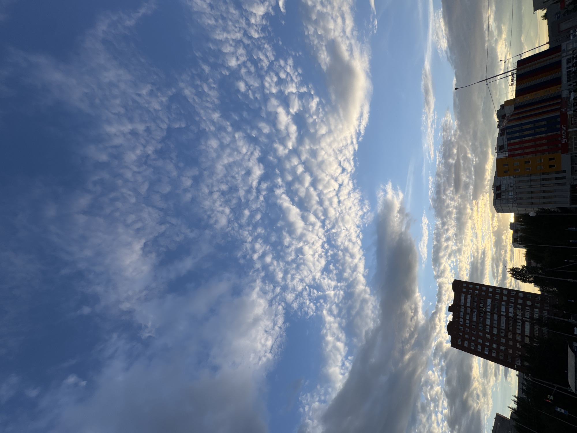 A low-angle, wide shot captures a vibrant sky filled with scattered, cumulus clouds against a bright blue expanse. Sunlight filters through the clouds, creating a soft, ethereal glow. In the foreground, silhouetted buildings and trees stand against the illuminated sky. To the left, a tall, dark-red apartment building with numerous windows dominates the frame, tilted slightly to the right. To the right, a lower, more modern building features a striking facade of red, yellow, and blue vertical stripes, with the word "МАГНИТ" visible on its side. Streetlights and utility wires crisscross the lower portion of the image, indicating an urban environment. The overall impression is one of a city skyline at dusk or dawn, bathed in the warm light of the setting or rising sun.