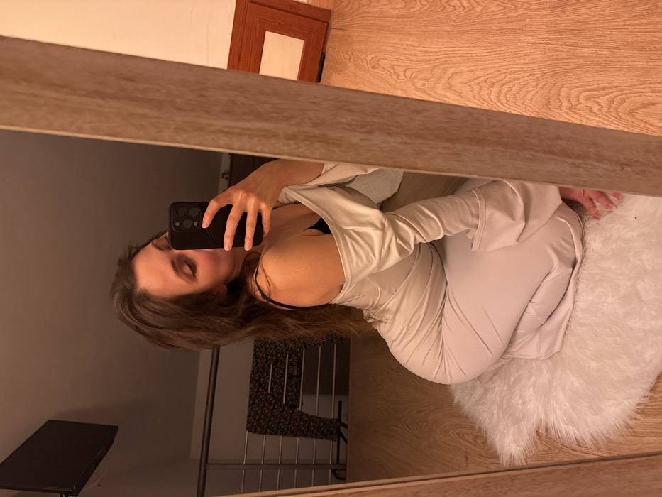 An image taken in Cava de' Tirreni, Italy, depicts a female individual captured in a mirror selfie within an indoor setting. The individual, possessing long brown hair, is shown in a kneeling or crouched position on a white, fluffy rug. She is wearing a light-colored, possibly beige or off-white, long-sleeved garment with an off-the-shoulder design, revealing a black bra strap on her left side. Her left hand holds a black smartphone, equipped with a pop socket, positioned to take the photograph.
The mirror, framed in light wood, reflects the interior of what appears to be a private residence. Visible in the background reflection are a black flat-screen television mounted high on a wall, a metal railing (suggesting a loft or upper level), and a dark patterned garment draped over the railing. To the right, a brown wooden picture frame leans against a wall on a light wood-grain floor. The overall lighting is warm-toned.