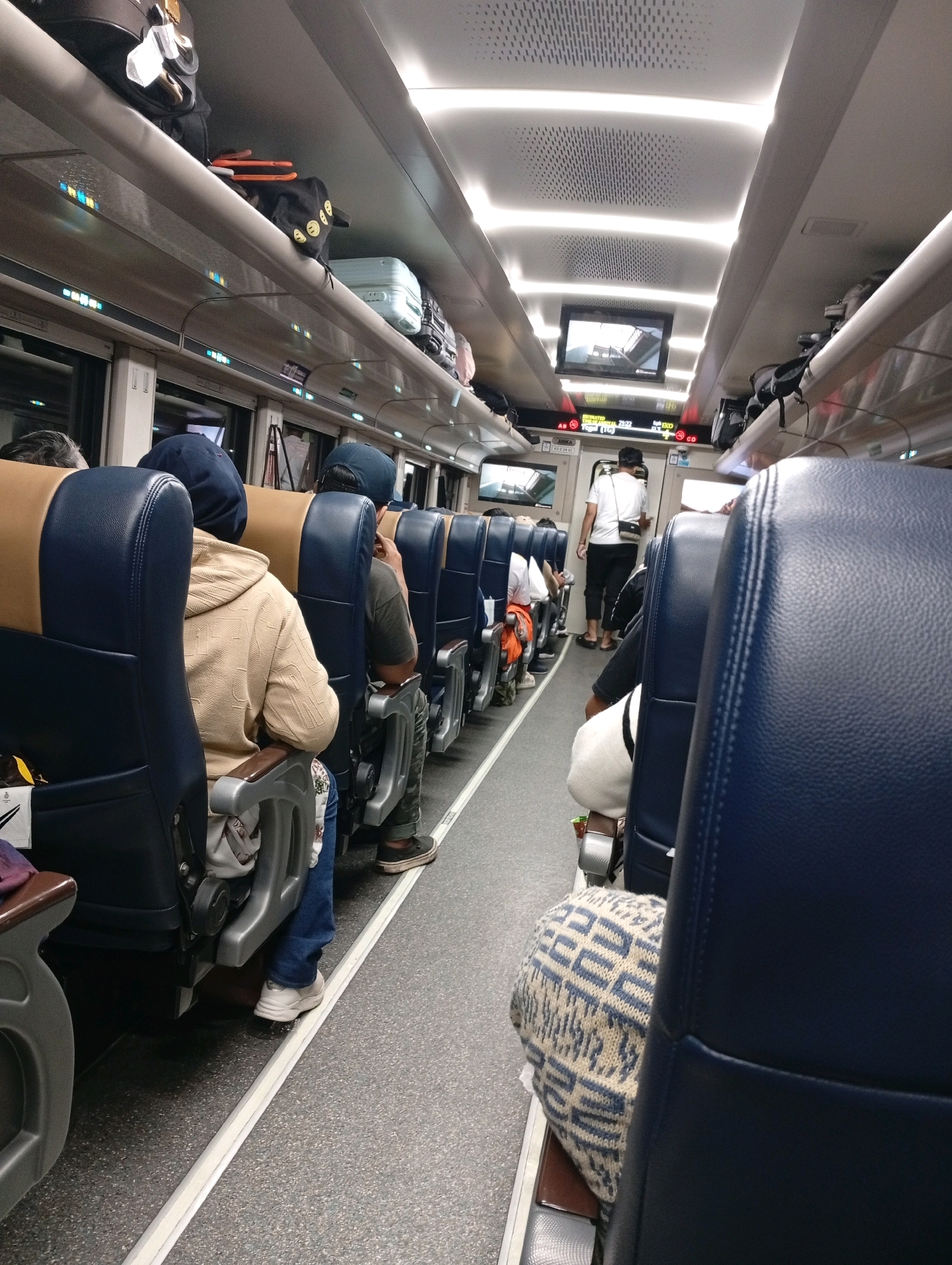 The image depicts the interior of a train car, likely at a station in Cirebon, Indonesia. Rows of blue and brown upholstered seats line the aisle. Several passengers are visible, mostly from the back, seated and facing forward. Luggage is stowed on overhead racks. A screen displays information above the aisle. The train appears to be either boarding or disembarking passengers, given the presence of individuals standing and walking in the aisle. The lighting is artificial, with LED strips running along the ceiling.