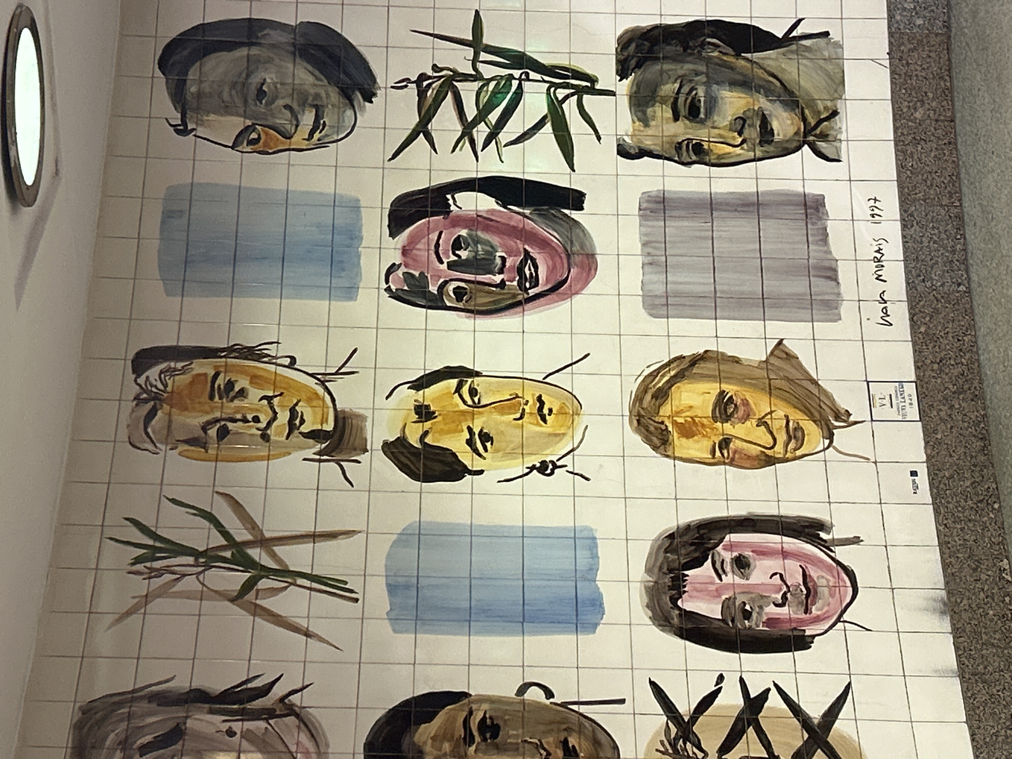 This image shows a mural made of tiled panels depicting a series of faces and abstract vegetal designs. The faces are drawn in a loose, expressive style with visible brushstrokes. Each panel features a different artistic rendering, using different color palettes and line work. Some tiles feature abstract blocks of color in blue and gray. Some tiles also have botanical depictions, such as stylized leaves and branches. Text "hola morais 1997" and "V-L VIUVA LAMERO 1849" are located on the right of the mural. The location is Unknown, Unknown.
