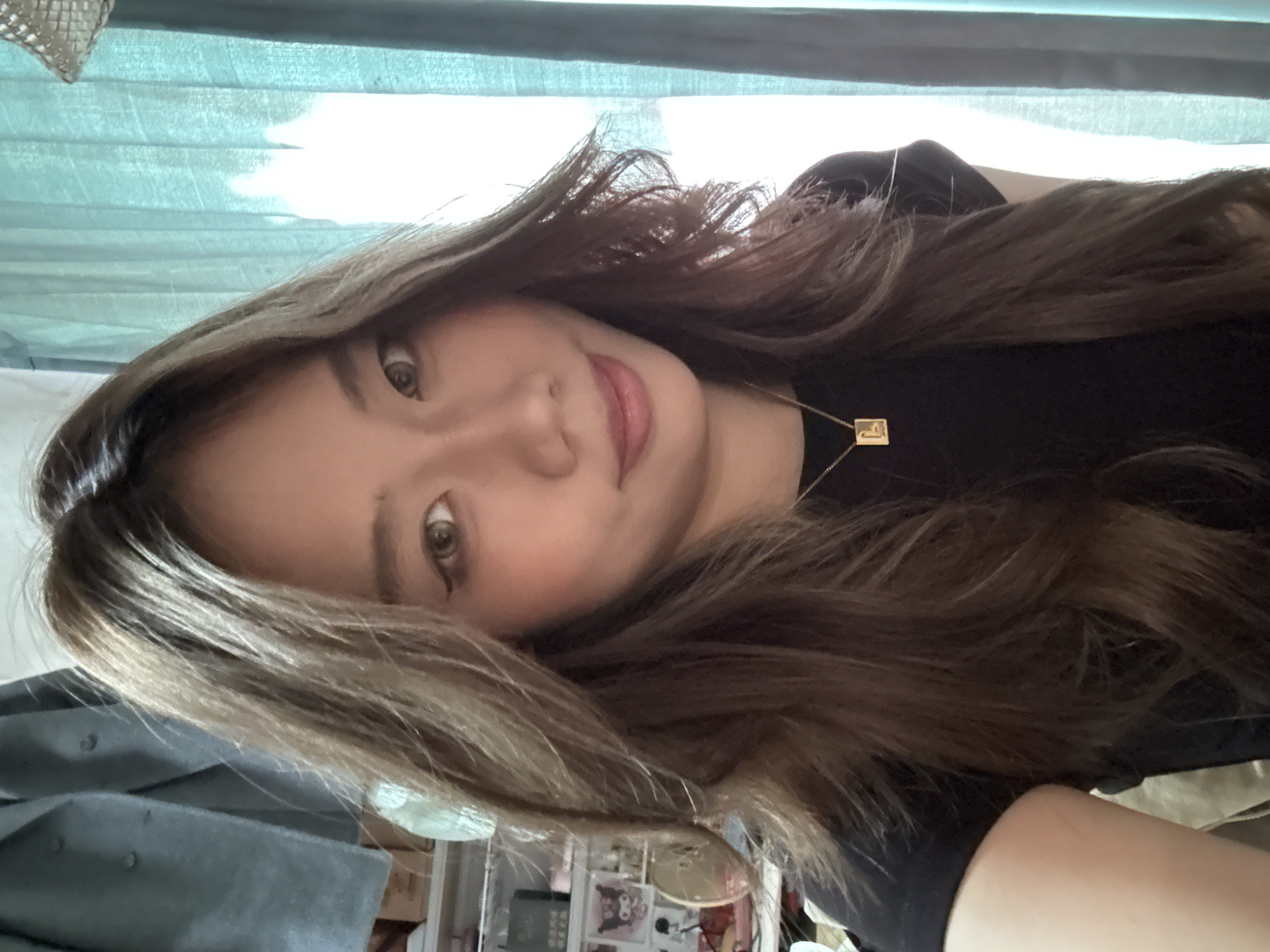 A young woman with long brown hair and fair skin is looking at the camera. She is wearing a black t-shirt and a gold necklace. The background is a room with a blue curtain and a shelf with various items. The location in the city of Nan'an District, China, cannot be confirmed from the provided image.