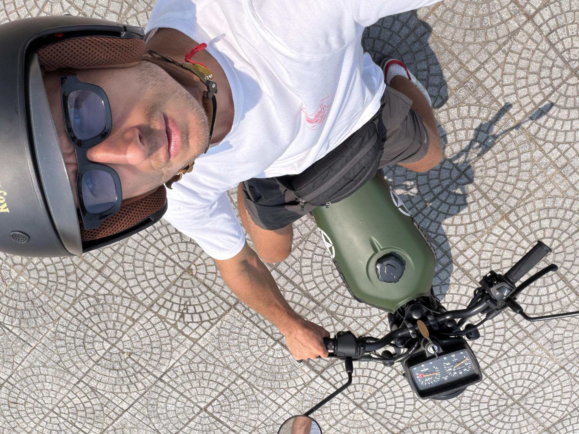 A high-angle, full shot of a man on a green motorcycle on a patterned, grey-tiled surface. The man wears a black helmet with brown lining, sunglasses, a white t-shirt, and dark shorts with a black fanny pack. He is holding the motorcycle handlebars. A side mirror is visible on the lower left. The motorcycle features a black dashboard with gauges. The location is suggested to be Đà Nẵng, Vietnam. The background is the patterned grey tiling and the upper half of the man's body. The light source appears to be from above and slightly behind the subject, casting shadows on the pavement.