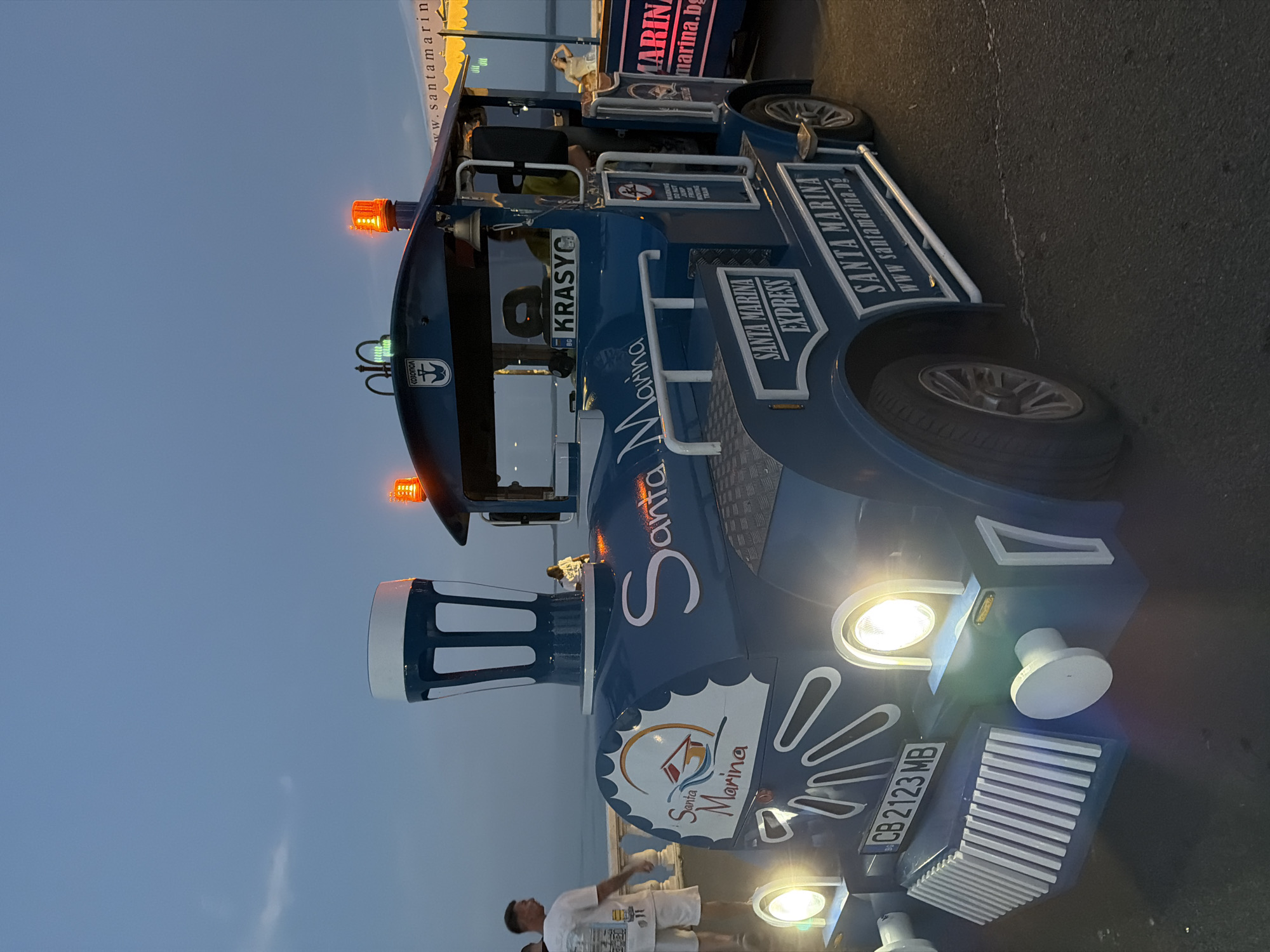 A blue tourist train labeled "Santa Marina Express" is parked on asphalt near a body of water, likely the sea, at dusk. The train has a stylized locomotive front with large headlights, a grill, and a chimney-like structure. Orange flashing lights are visible on top of the train's cab. A Bulgarian license plate reading "CB 2123 MB" is attached to the front. The train's side is decorated with the "Santa Marina" logo and website address. A person wearing a white t-shirt and shorts stands to the left of the train, facing away from the camera. Another person is visible inside the train's cab. In the background, a person is standing near a yellow awning. The sky is a dusky blue. The scene appears to be on the Burgas Sea Garden promenade, a popular public space in the city of Burgas, Bulgaria, known for its recreational activities and sea views.