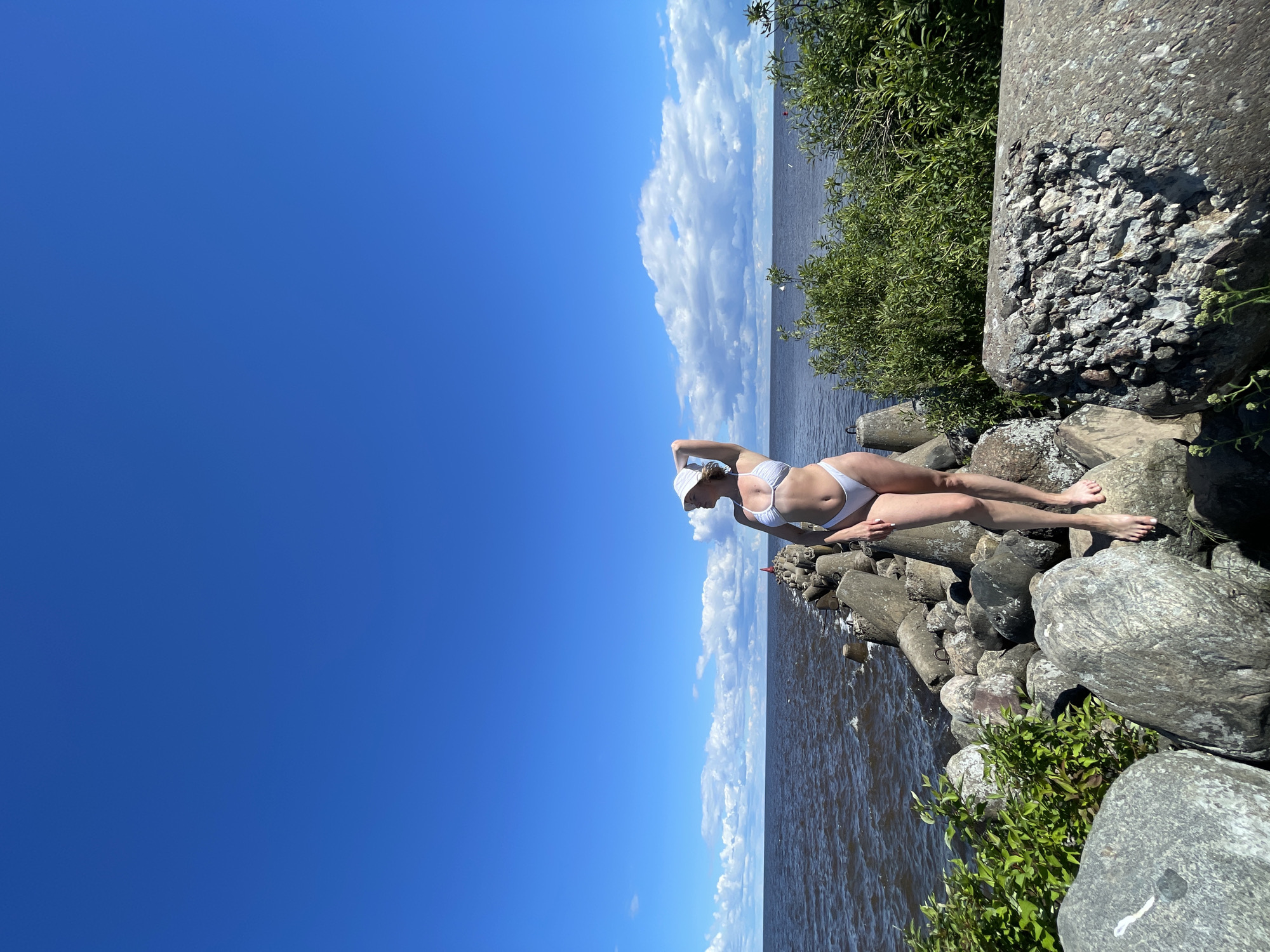 A woman in a white bikini and a white bucket hat stands on a rocky breakwater under a bright blue sky with scattered clouds. The breakwater extends into dark, choppy water. Green foliage grows on the sides of the breakwater. The woman is facing away from the camera, with her right arm raised, touching her hat. The scene appears to be a coastal area.